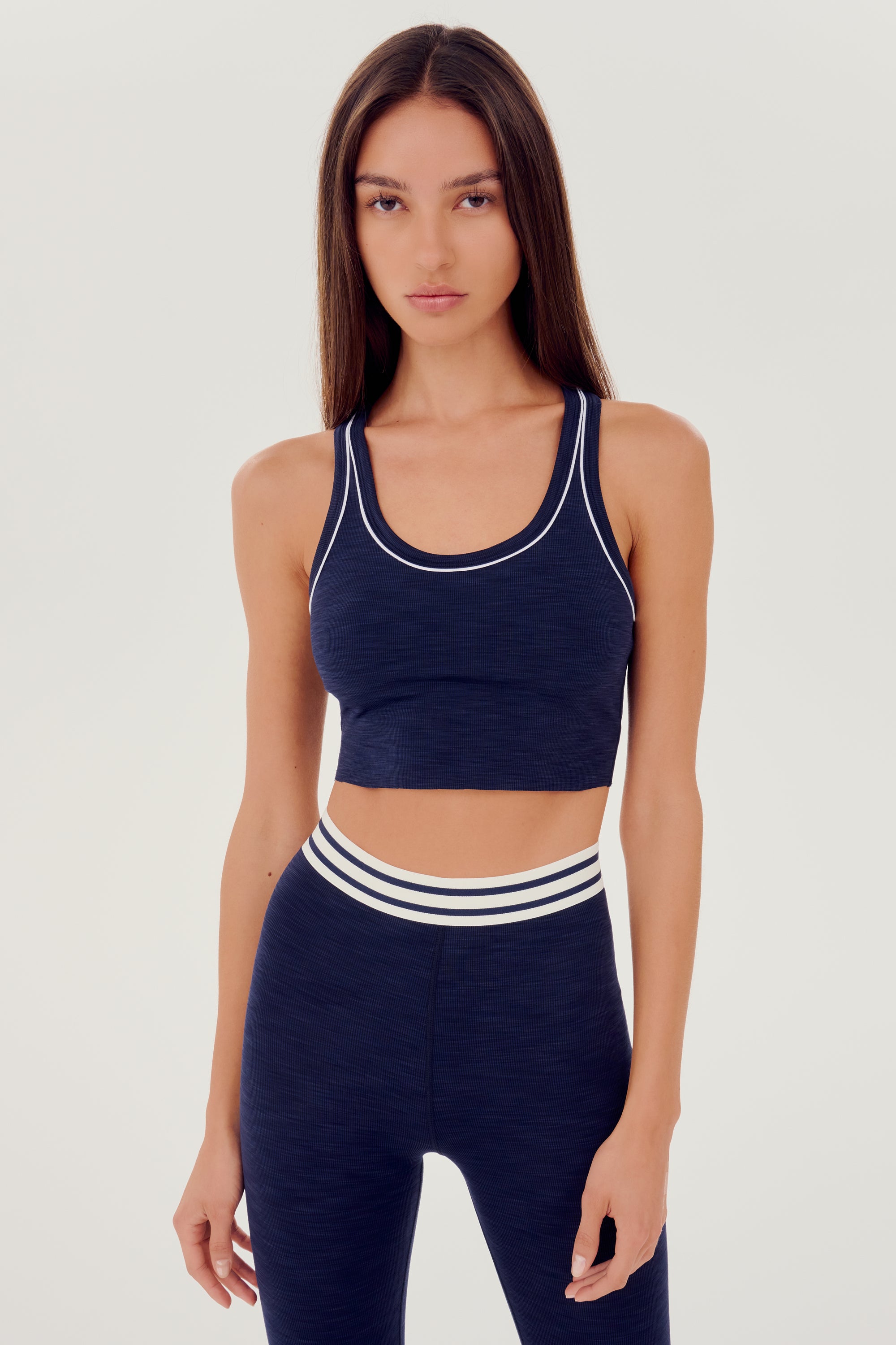 Front view of girl wearing a dark blue ribbed bra that stops at the ribs with thin white line along arms and collar with blue leggings with white and blue waistband 