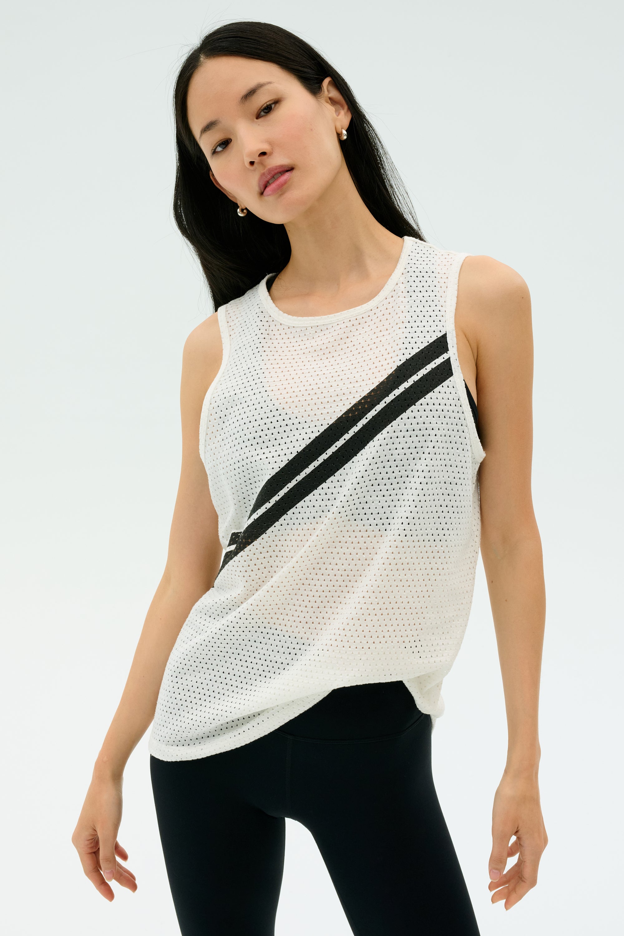 A woman models the SPLITS59 Bandit Mesh Tank in white/black, a sleeveless mesh top with two diagonal black stripes, paired with black leggings against a light background.
