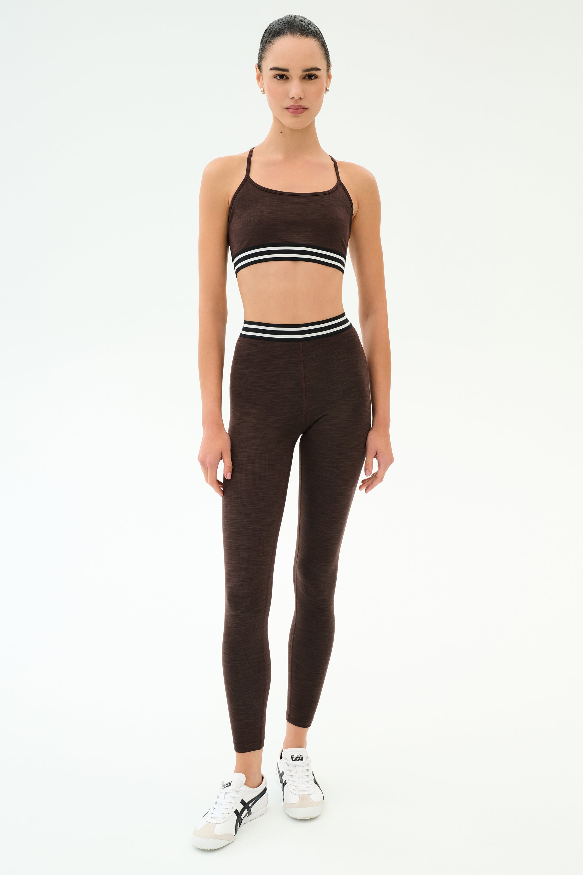 A woman wears the SPLITS59 Cali Active Rib Bra in Heather Dark Chocolate with matching leggings, featuring white and black striped waistbands, standing against a plain white background.