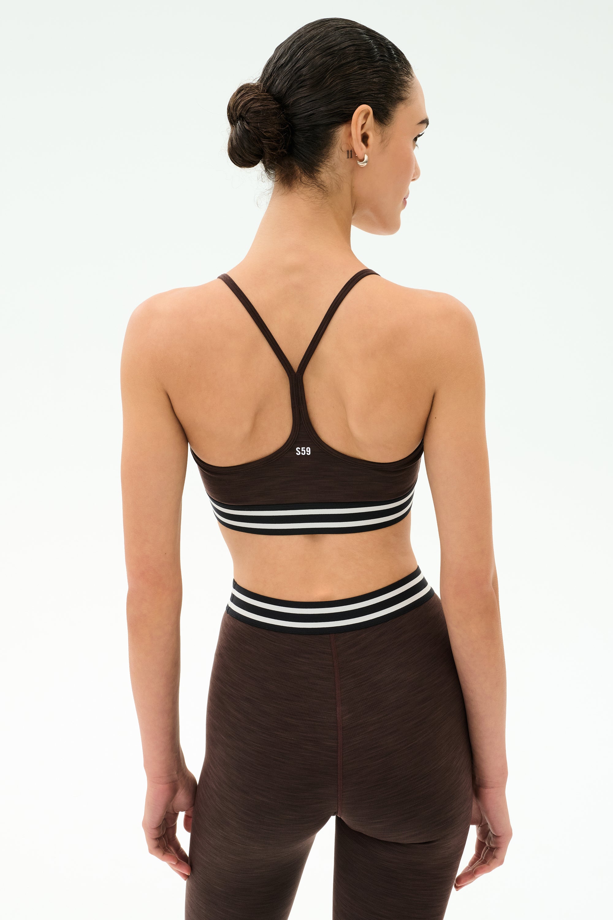 A woman stands with her back to the camera, wearing the SPLITS59 Cali Active Rib Bra in Heather Dark Chocolate and matching leggings with white and black striped elastic bands, set against a plain background.