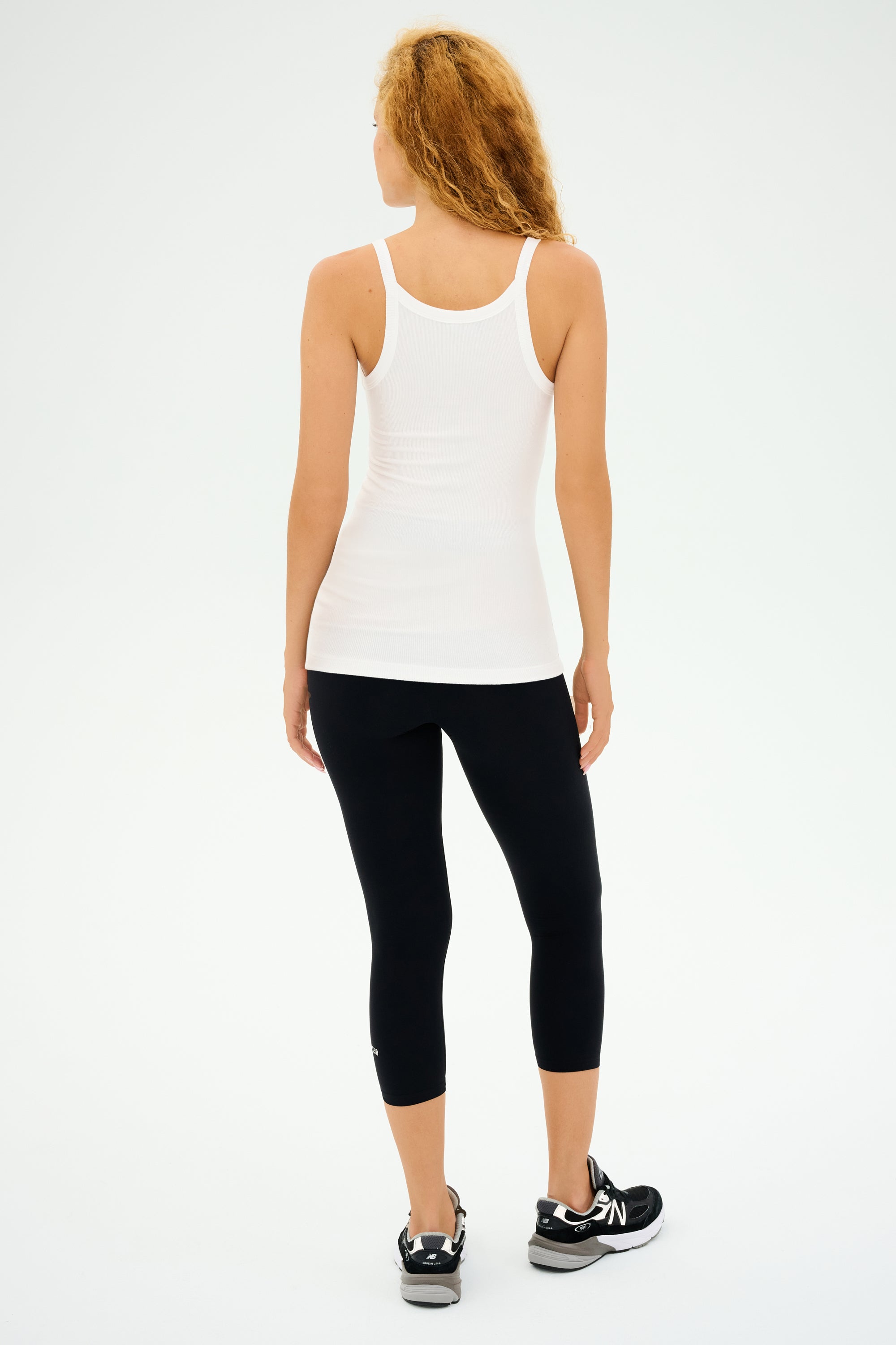 A person with long curly hair wears the Charlie Rib Tank in white by SPLITS59, paired with black leggings and sneakers, standing confidently against a white background.