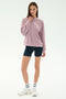 A woman stands against a plain background wearing the SPLITS59 Joy Velour Crewneck in Blush, black spandex bike shorts, white sneakers, and white socks with navy stripes.