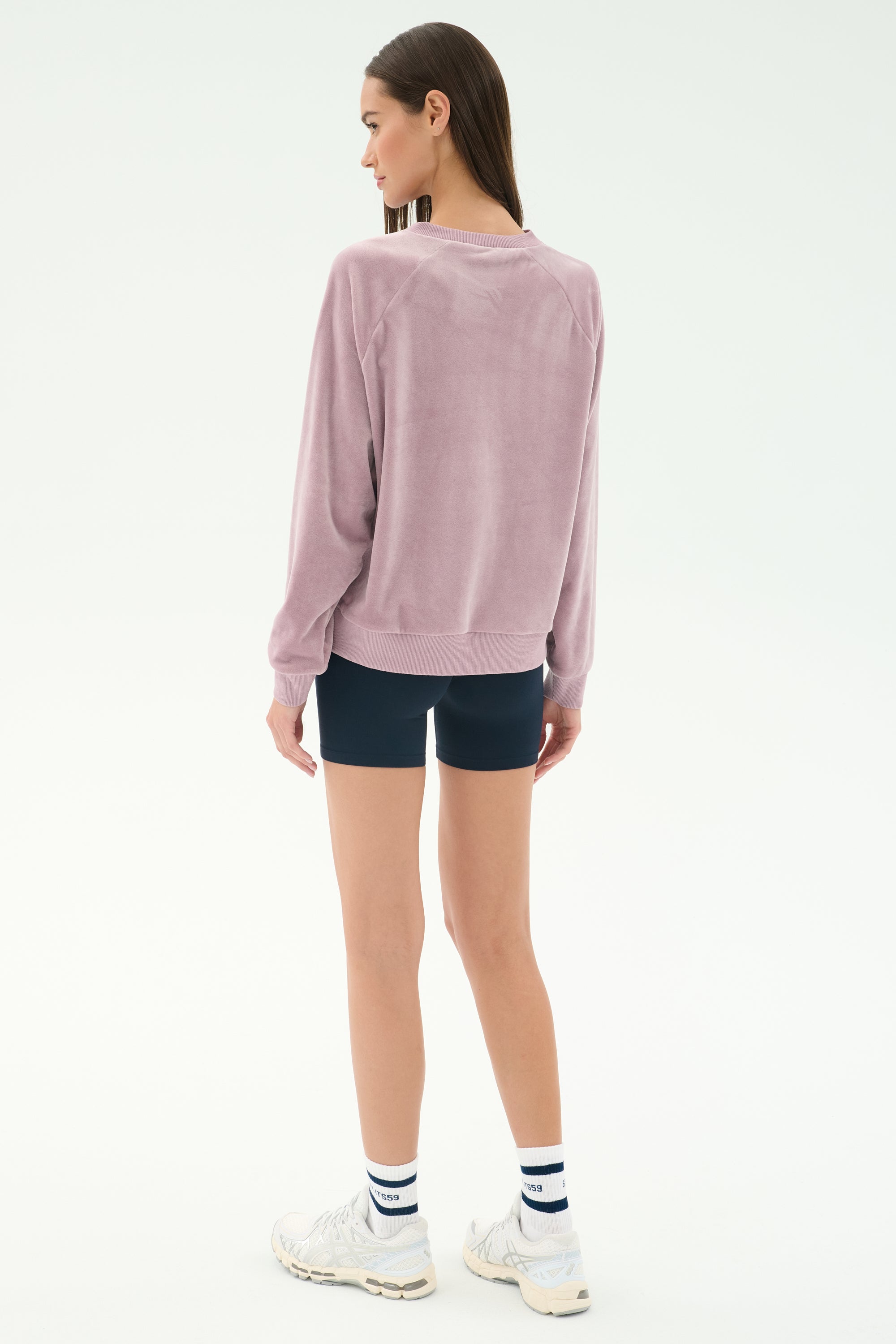 A woman faces away, wearing the SPLITS59 Joy Velour Crewneck in Blush, navy shorts, white athletic shoes, and white socks with navy stripes against a plain light background.