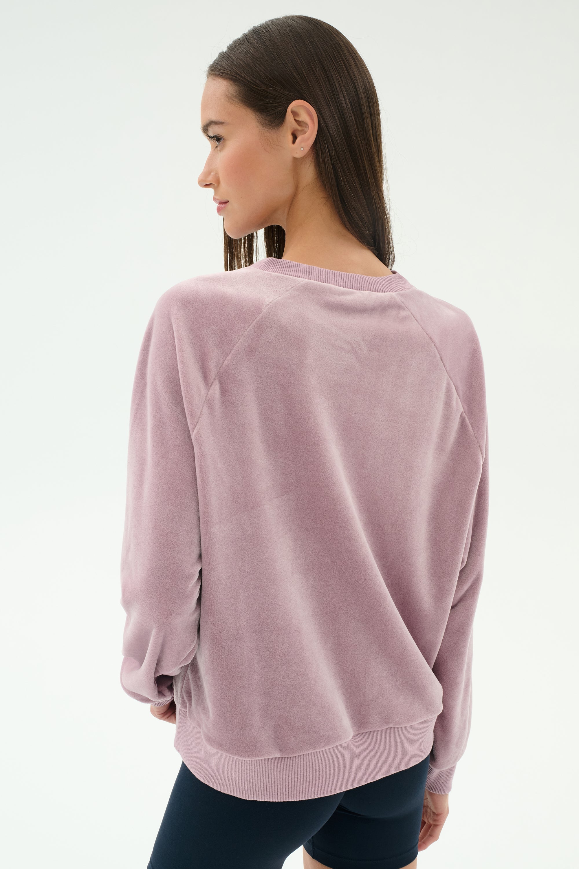 A woman with straight brown hair wears the SPLITS59 Joy Velour Crewneck in blush and black spandex leggings, standing against a plain white background.