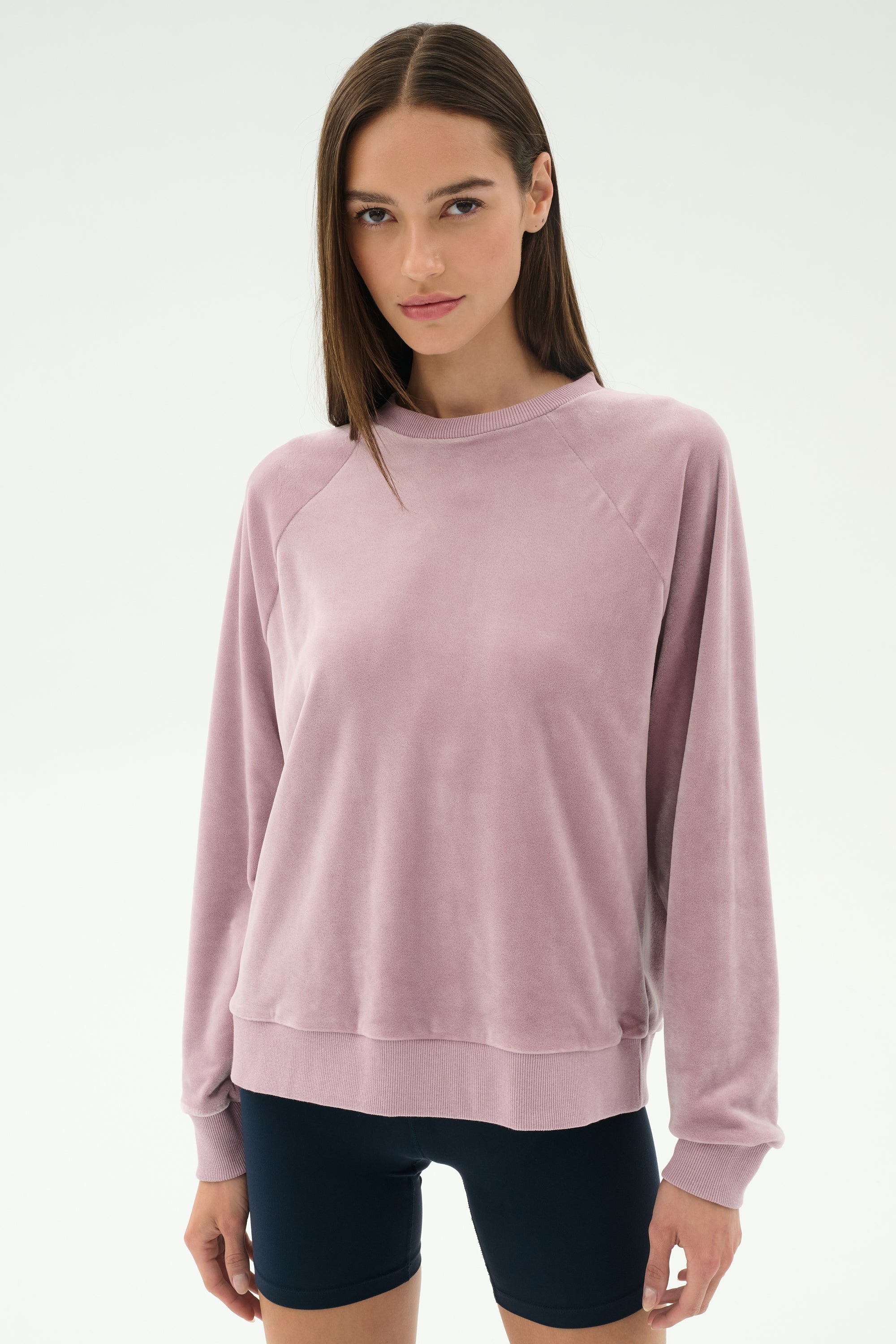 A woman stands against a plain background wearing the SPLITS59 Joy Velour Crewneck in Blush and black shorts, looking directly at the camera.