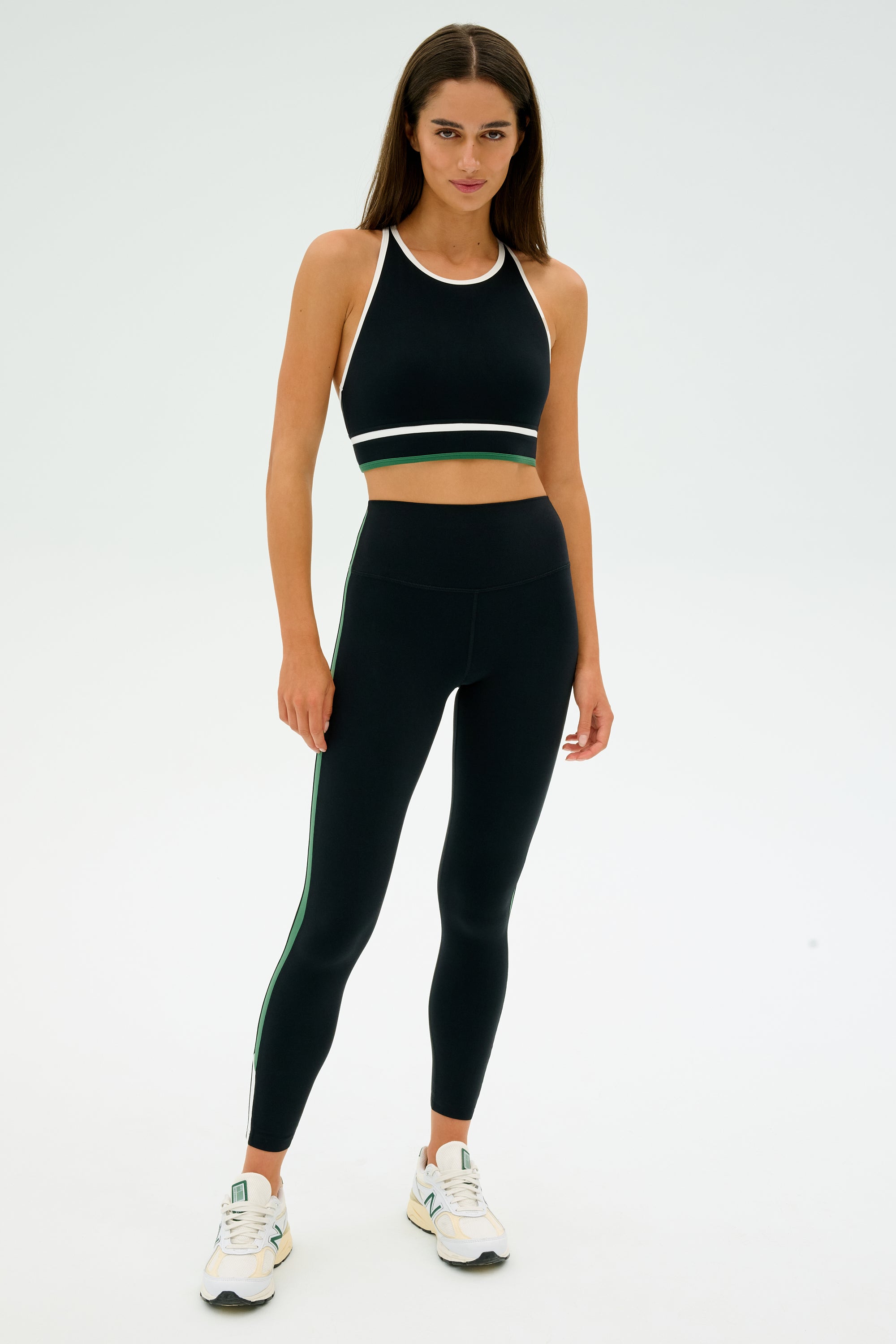 A person stands in a neutral pose against a plain background wearing the Layla Airweight Bra in Black/Arugula by SPLITS59, paired with matching athletic wear. They're wearing sneakers and looking directly at the camera.