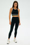 A person stands in a neutral pose against a plain background wearing the Layla Airweight Bra in Black/Arugula by SPLITS59, paired with matching athletic wear. They're wearing sneakers and looking directly at the camera.