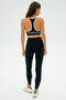 A person in the SPLITS59 Layla Airweight Bra - Black/Arugula and matching leggings stands with their back facing on a white background.