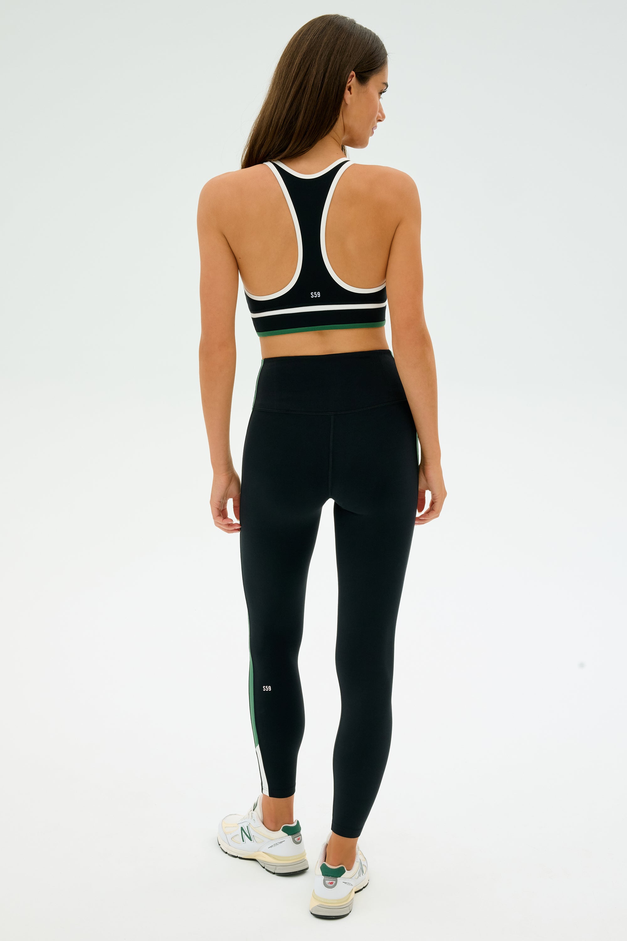 A woman in SPLITS59's sleek, black/white athletic ensemble featuring the stretchy Layla Airweight High Waist 7/8 leggings in Black/Arugula stands with her back to the camera, wearing a sports bra and sneakers against a plain backdrop.