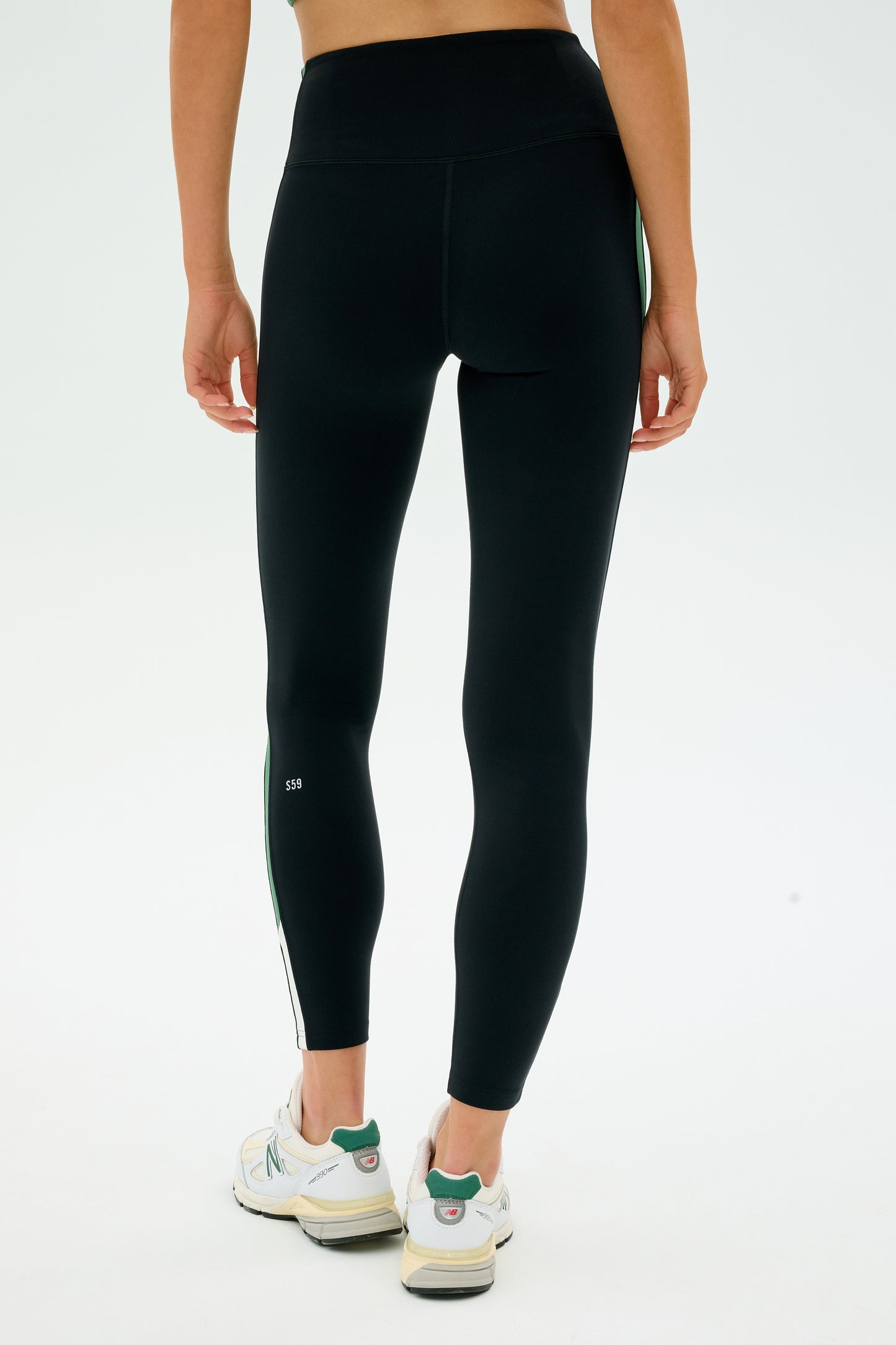 A person is wearing SPLITS59's Layla Airweight High Waist 7/8 Leggings in Black/Arugula and white sneakers, standing with their back to the camera against a plain background.