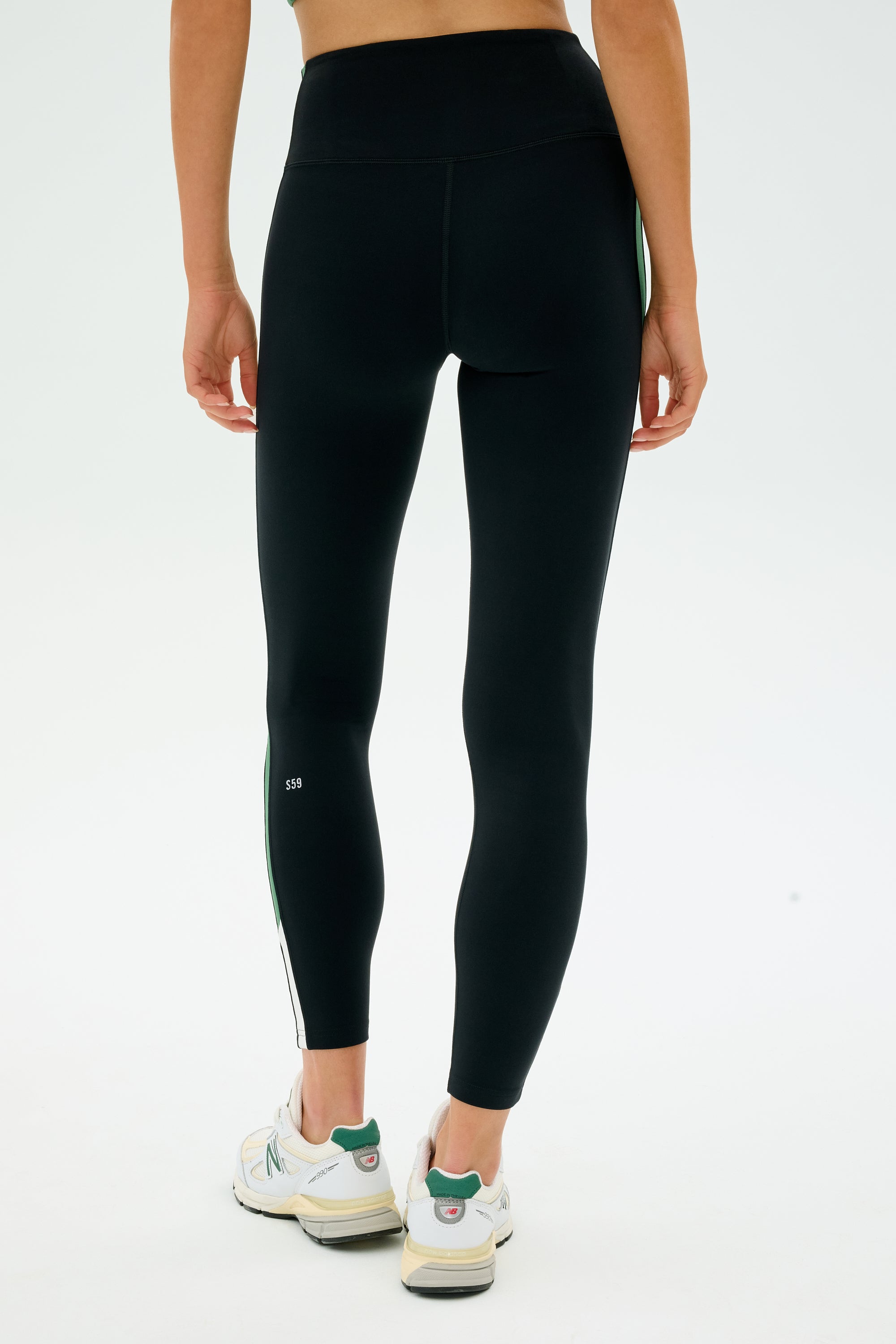 A person is wearing SPLITS59's Layla Airweight High Waist 7/8 Leggings in Black/Arugula and white sneakers, standing with their back to the camera against a plain background.