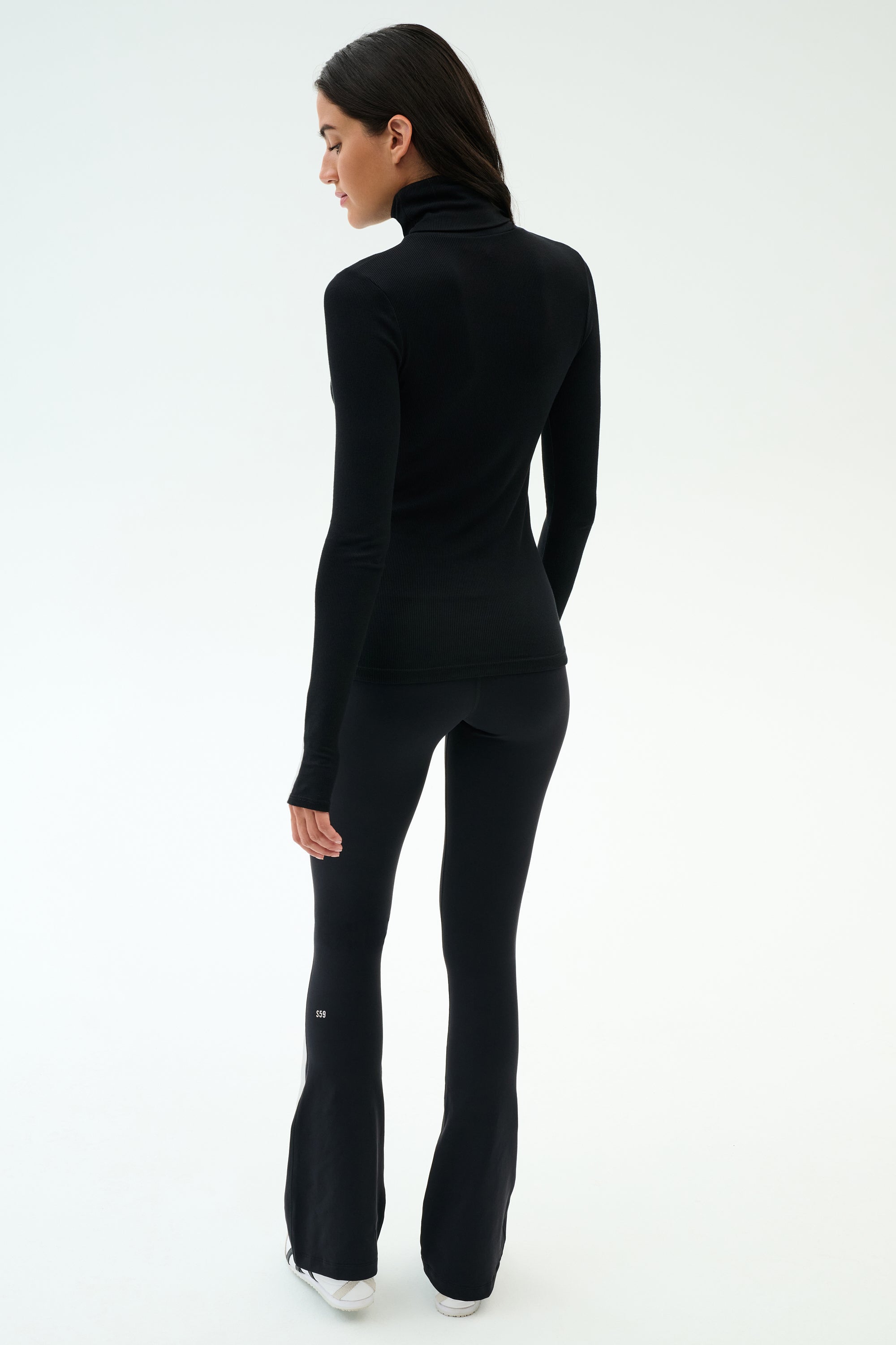 A woman stands facing away in SPLITS59’s Lennon Rib Turtleneck - Black/White and black flared pants with white sneakers—an effortlessly chic look for yoga or errands, set against a plain white background.