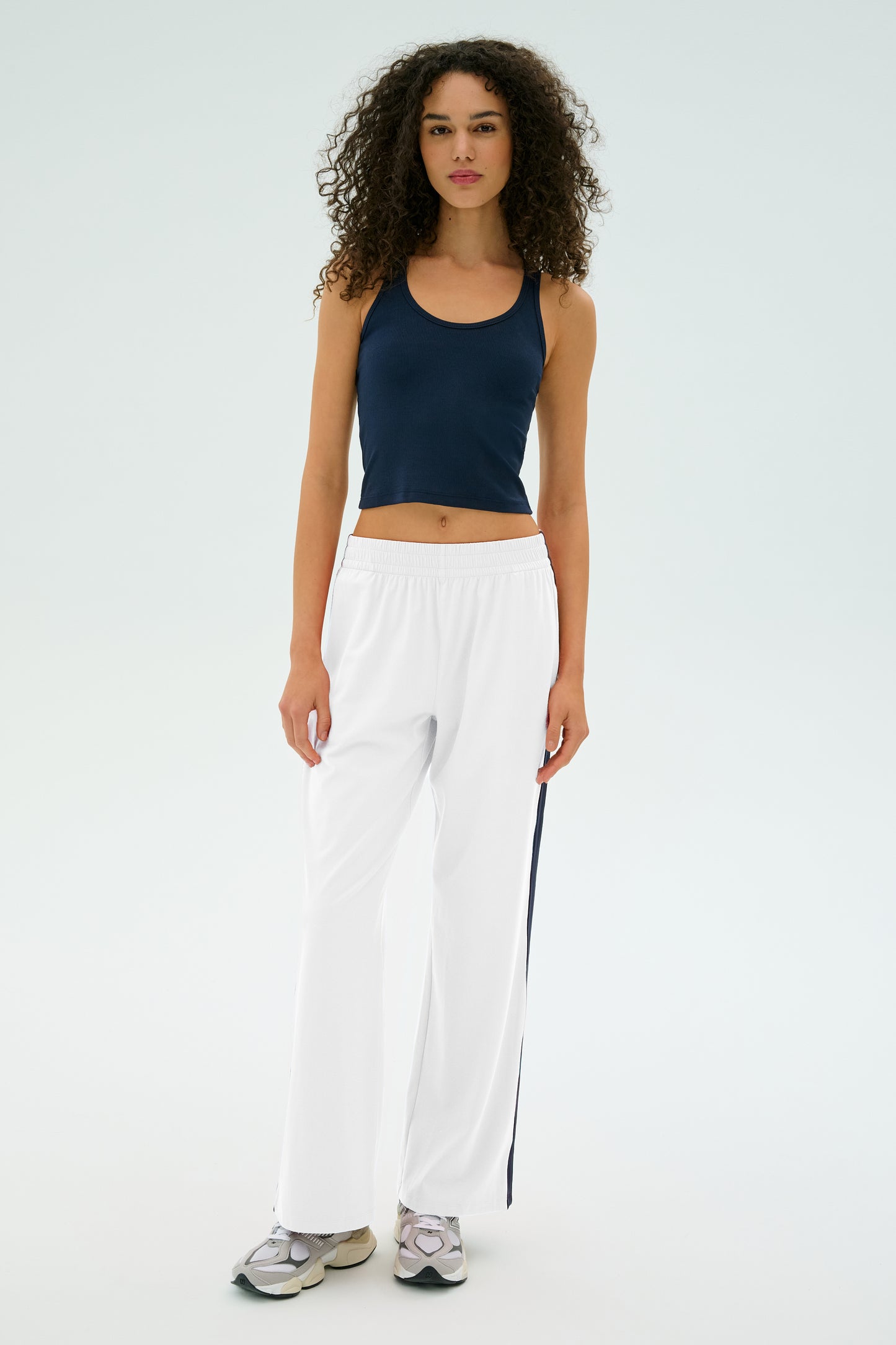 A person with curly hair is wearing a navy tank top, Maxie Rigor Track Pant by SPLITS59, and sneakers while standing against a light background.