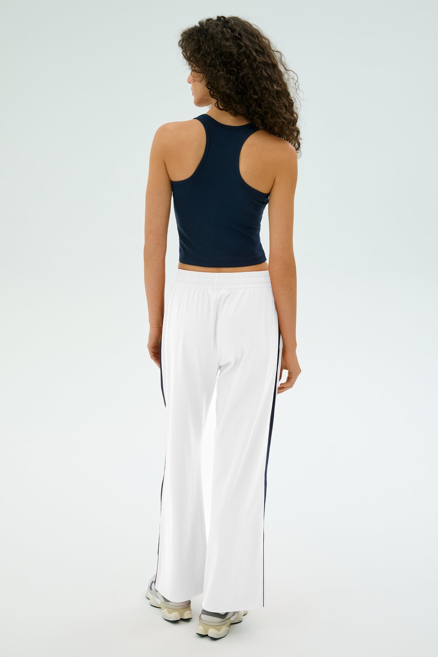 A person with curly hair, facing away, wears a navy tank top and SPLITS59's Maxie Rigor Track Pant in White/Indigo, featuring black side stripes, along with sneakers.