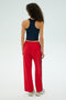With curly hair, a person dons a black tank top and vibrant Pirate Red/Indigo Maxie Rigor Track Pant by SPLITS59. They pair it with white sneakers, standing back to the camera against a plain backdrop for an effortlessly casual vibe.
