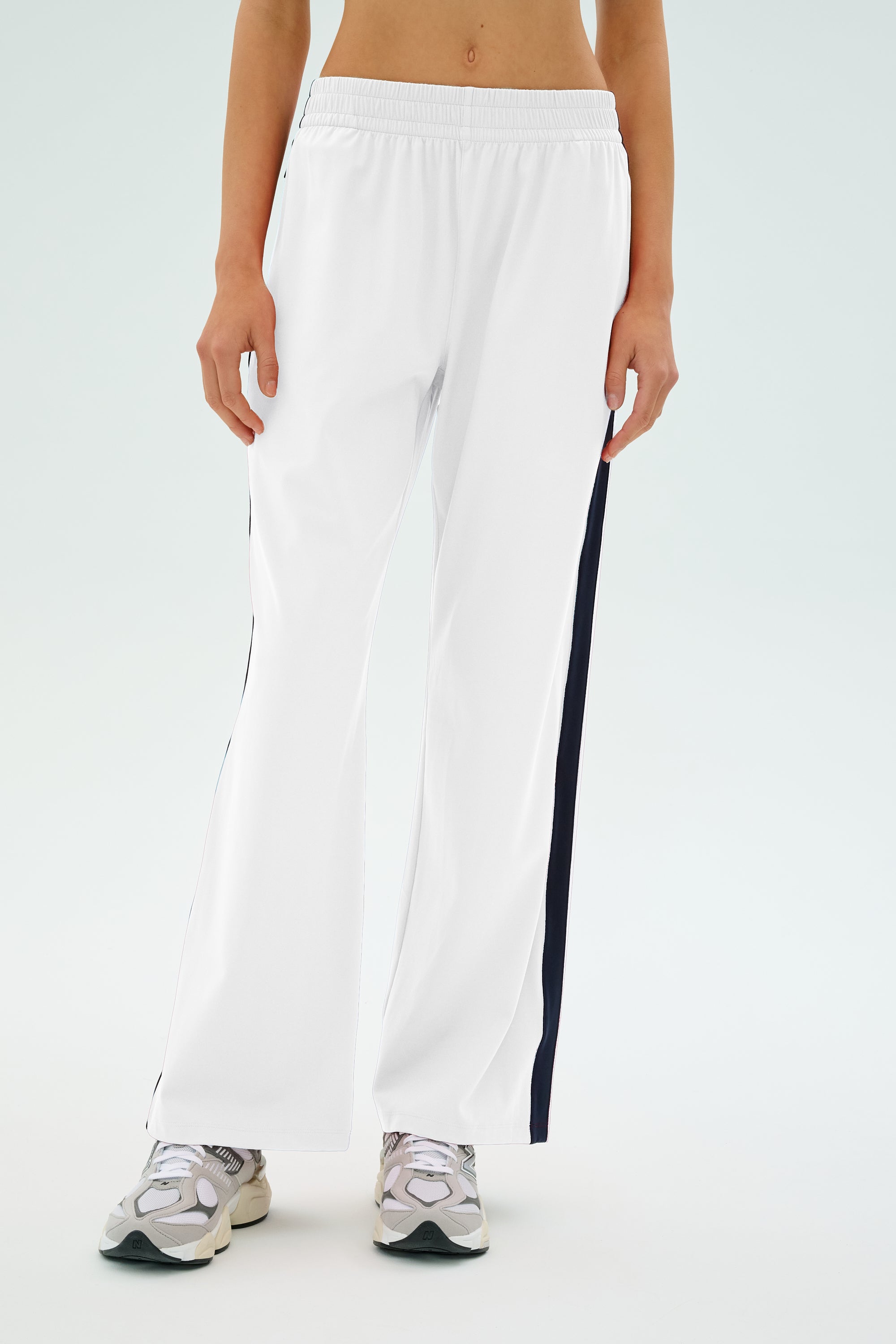 A person in SPLITS59's Maxie Rigor Track Pant in White/Indigo with black side stripes and white sneakers stands against a light background, exuding sporty elegance.
