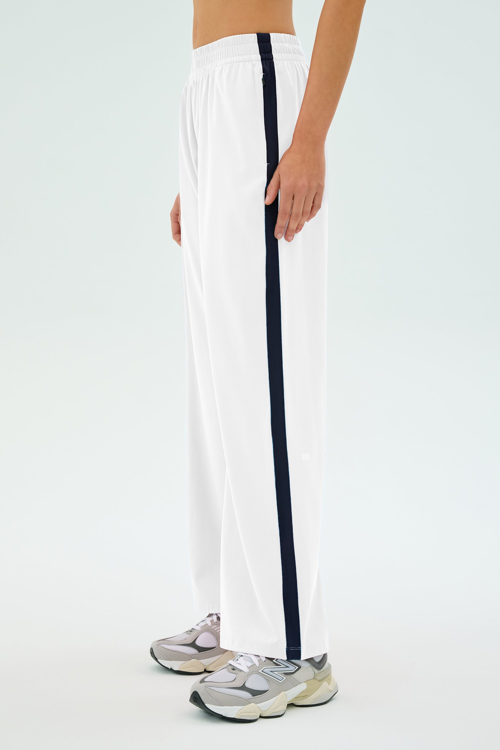 Wearing SPLITS59's Maxie Rigor Track Pant in White/Indigo with grey sneakers and a sleek track jacket, the individual stands against a light background.