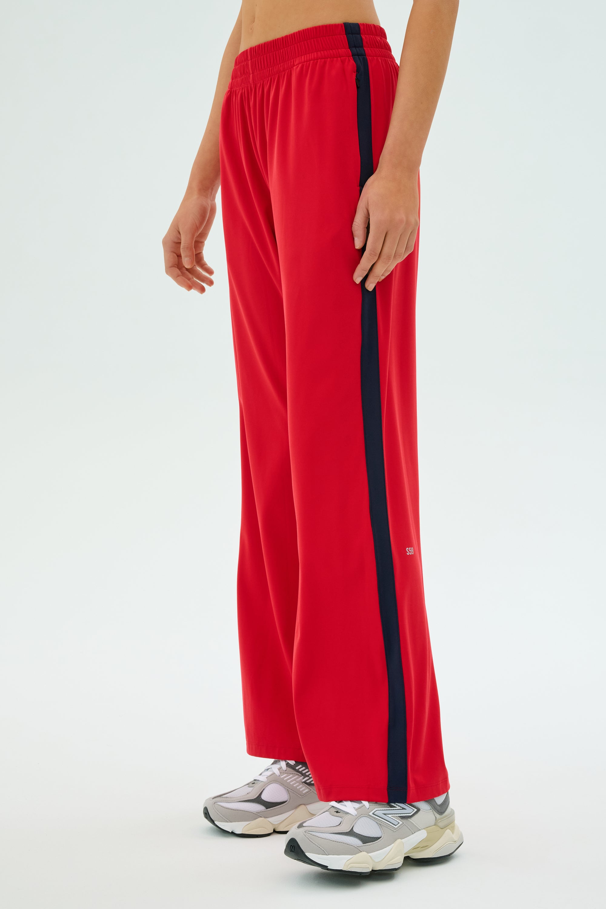 Against a plain background, a person wears SPLITS59's Maxie Rigor Track Pant in Pirate Red/Indigo with a sleek black stripe, paired with white sneakers for a fresh look.