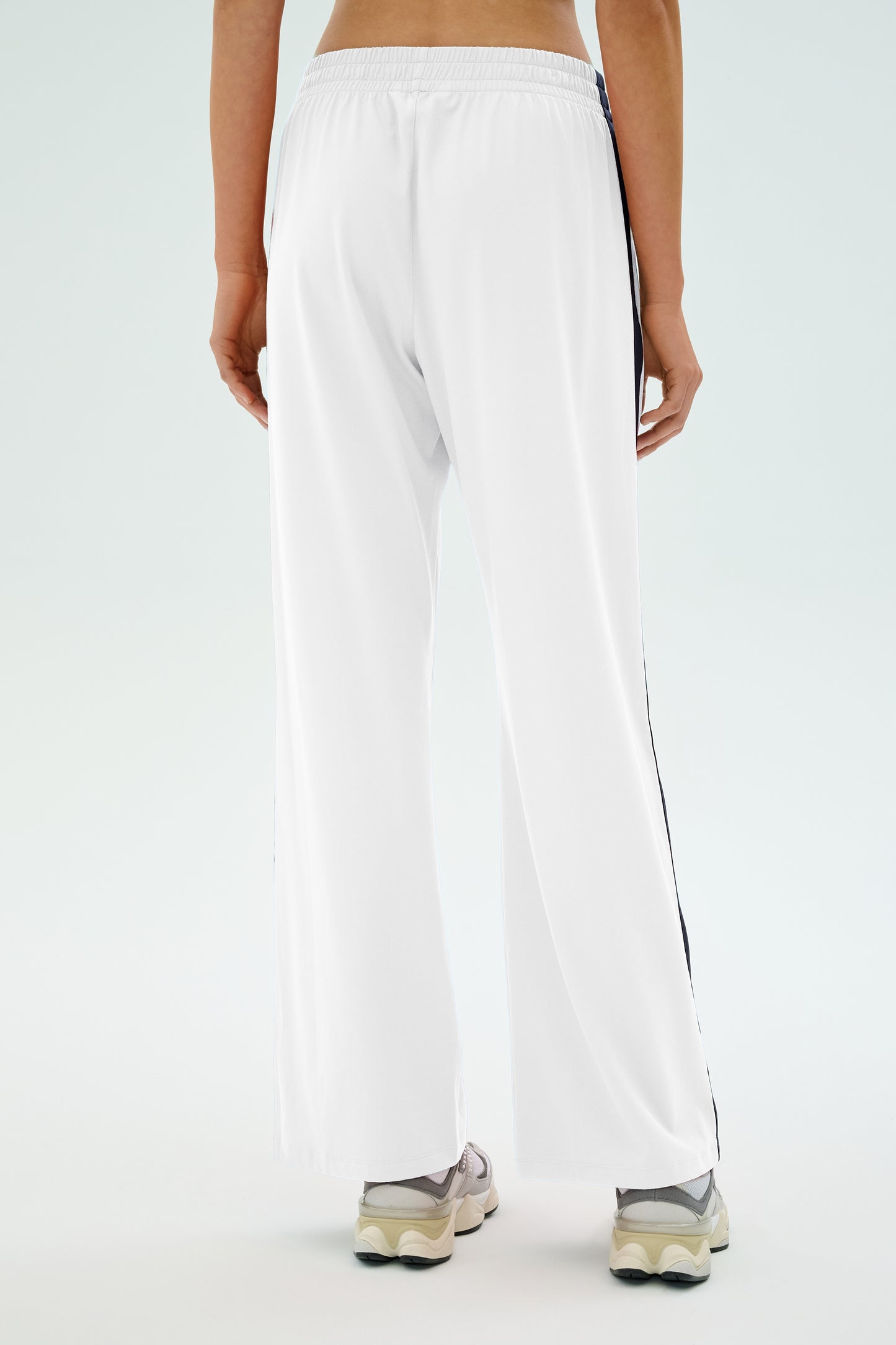 The person is seen from the back wearing SPLITS59's Maxie Rigor Track Pant in white with indigo side stripes. Paired with white sneakers on a plain background, these wide-leg pants provide comfort and style with their innovative Rigor fabric.