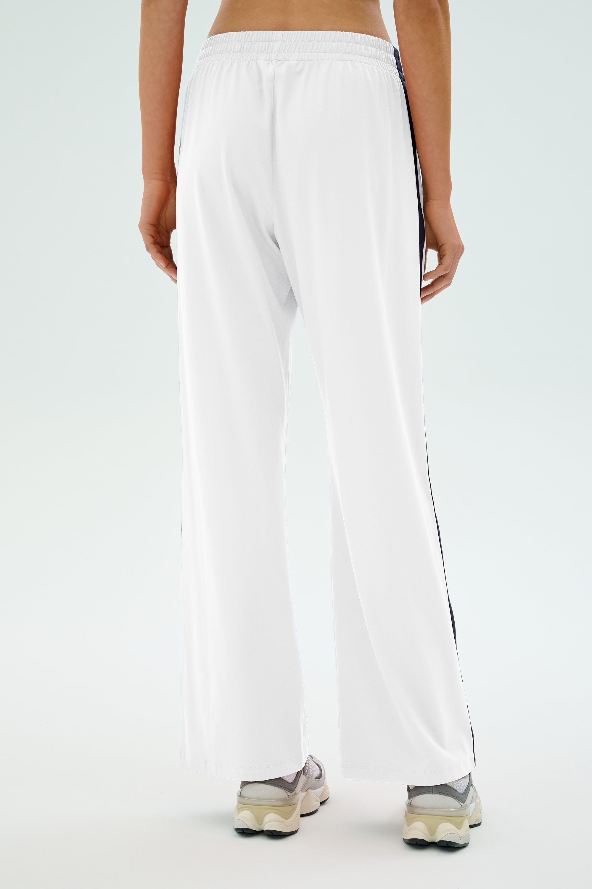The person is seen from the back wearing SPLITS59's Maxie Rigor Track Pant in white with indigo side stripes. Paired with white sneakers on a plain background, these wide-leg pants provide comfort and style with their innovative Rigor fabric.