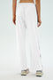 The person is seen from the back wearing SPLITS59's Maxie Rigor Track Pant in white with indigo side stripes. Paired with white sneakers on a plain background, these wide-leg pants provide comfort and style with their innovative Rigor fabric.