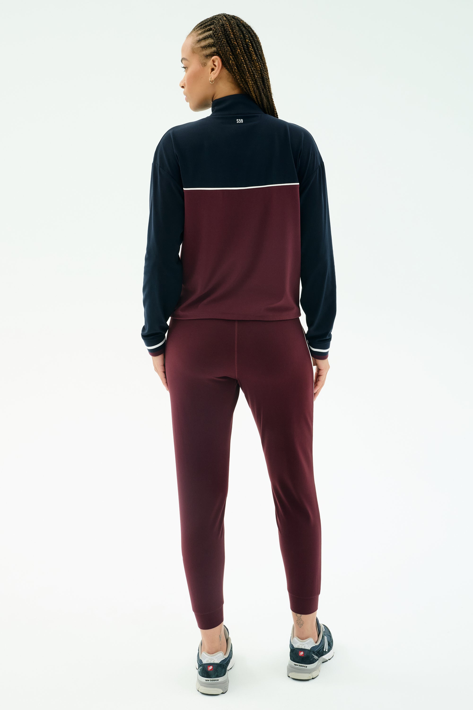 A person with braided hair faces away, wearing the SPLITS59 Millie Airweight Half Zip in Dark Cherry/Indigo and athletic shoes, shown against a plain white background.