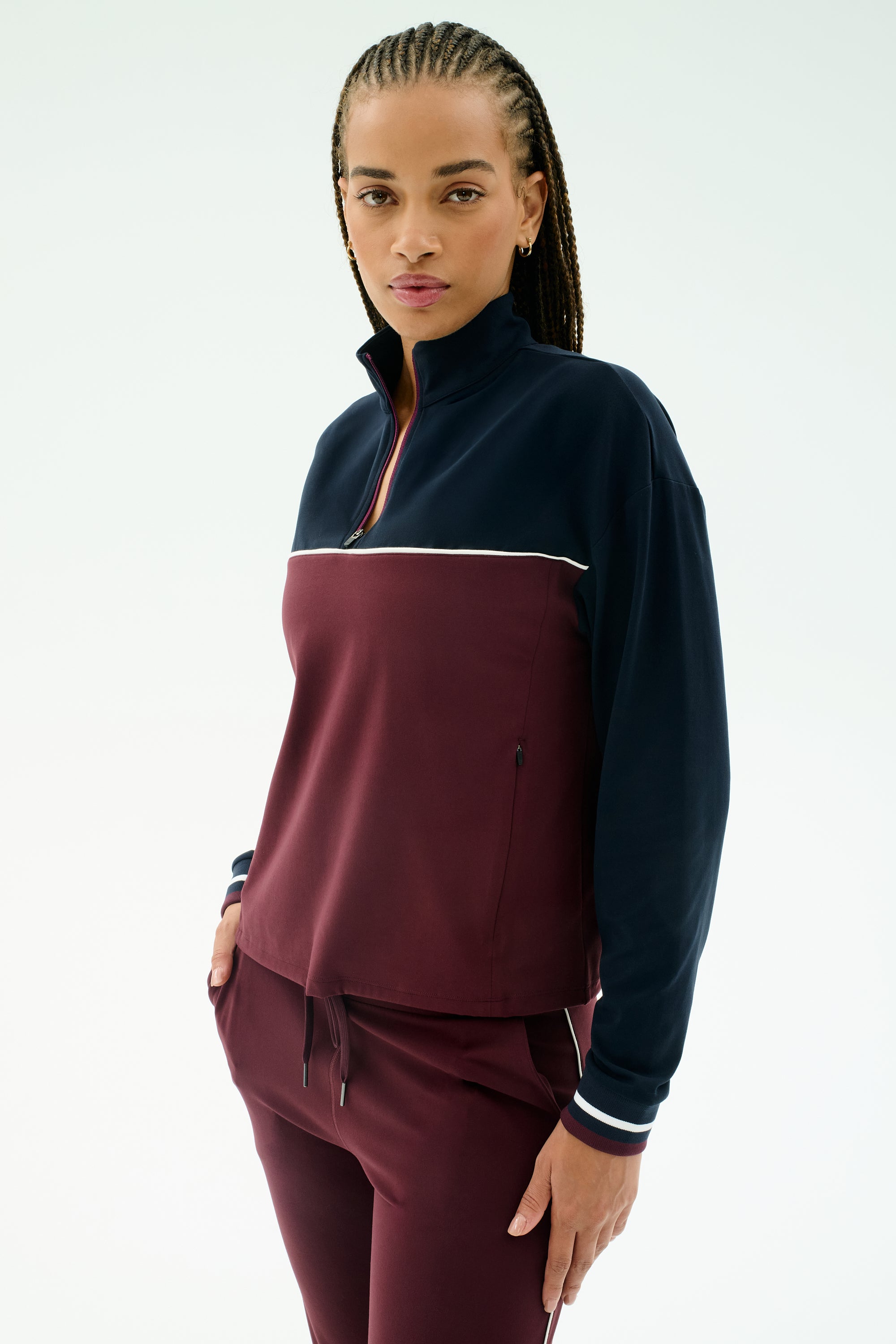 A woman wears the SPLITS59 Millie Airweight Half Zip in Dark Cherry/Indigo, a comfortable nylon-spandex blend track suit, standing with one hand in her pocket and looking at the camera against a plain white background.