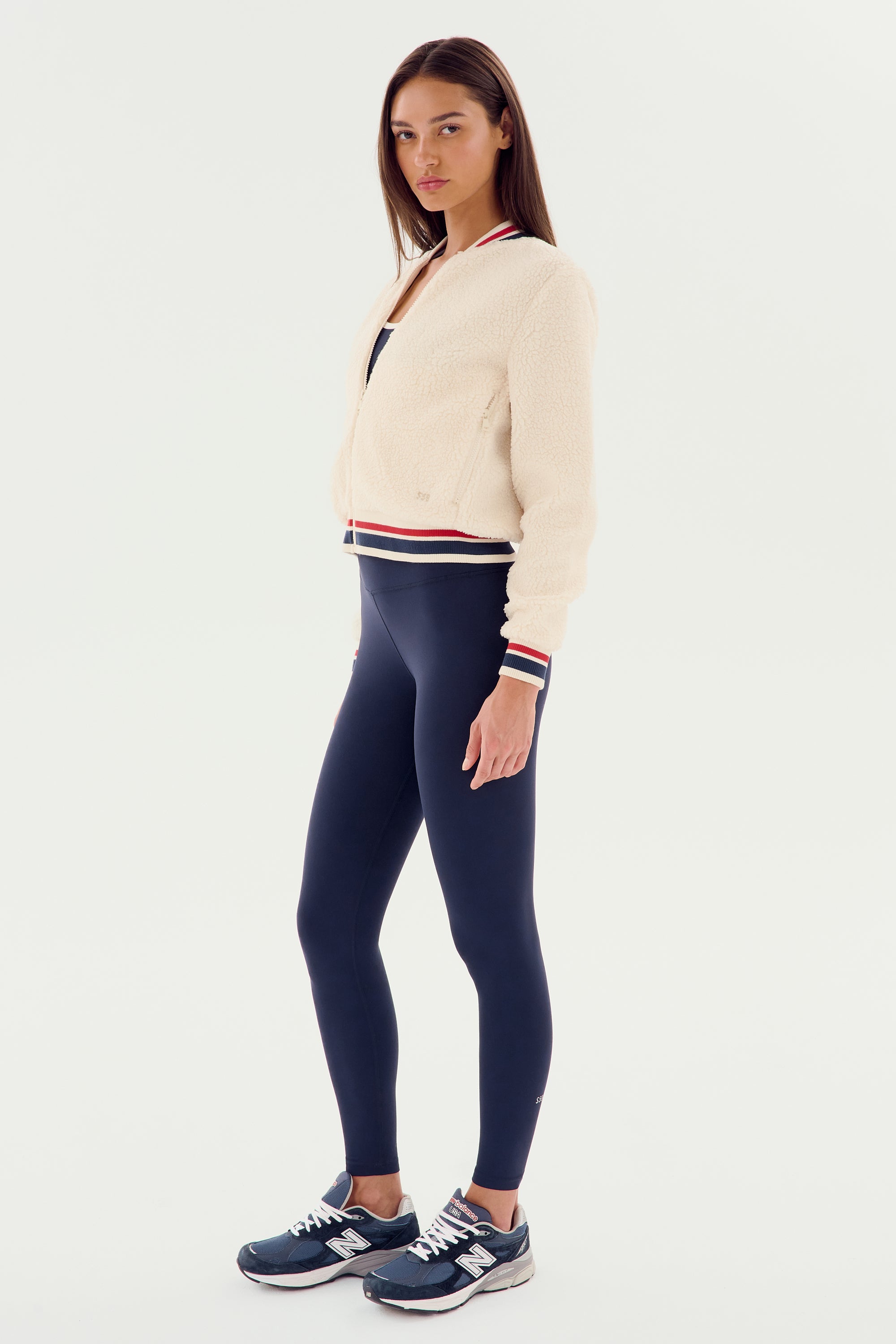 A woman stands against a white background wearing a cozy sportswear look: the SPLITS59 Willa Sherpa Jacket in cream, paired with navy leggings and navy sneakers.