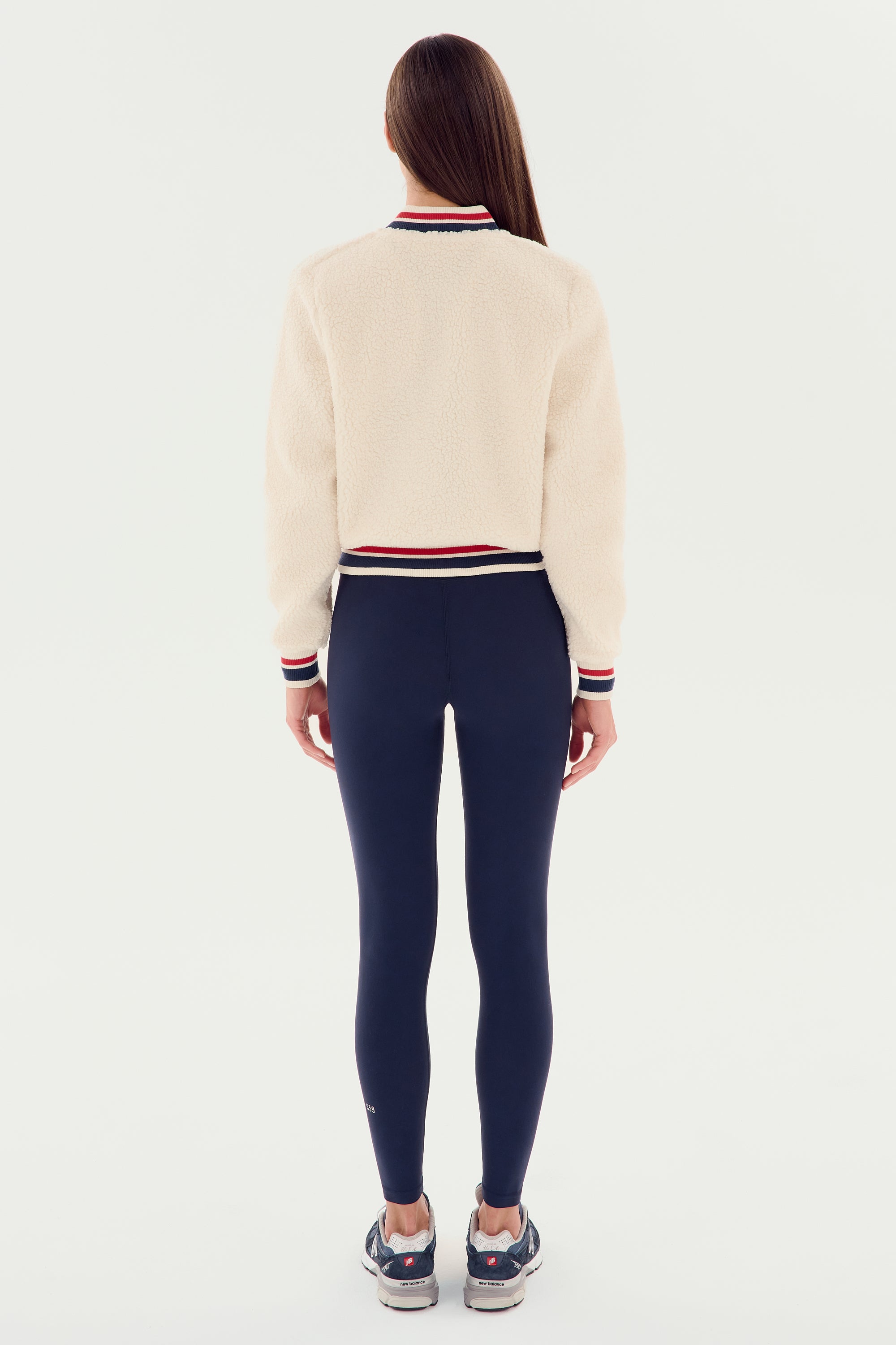 A woman stands facing away against a white background, wearing the SPLITS59 Willa Sherpa Jacket in cream, navy leggings, and gray sneakers.