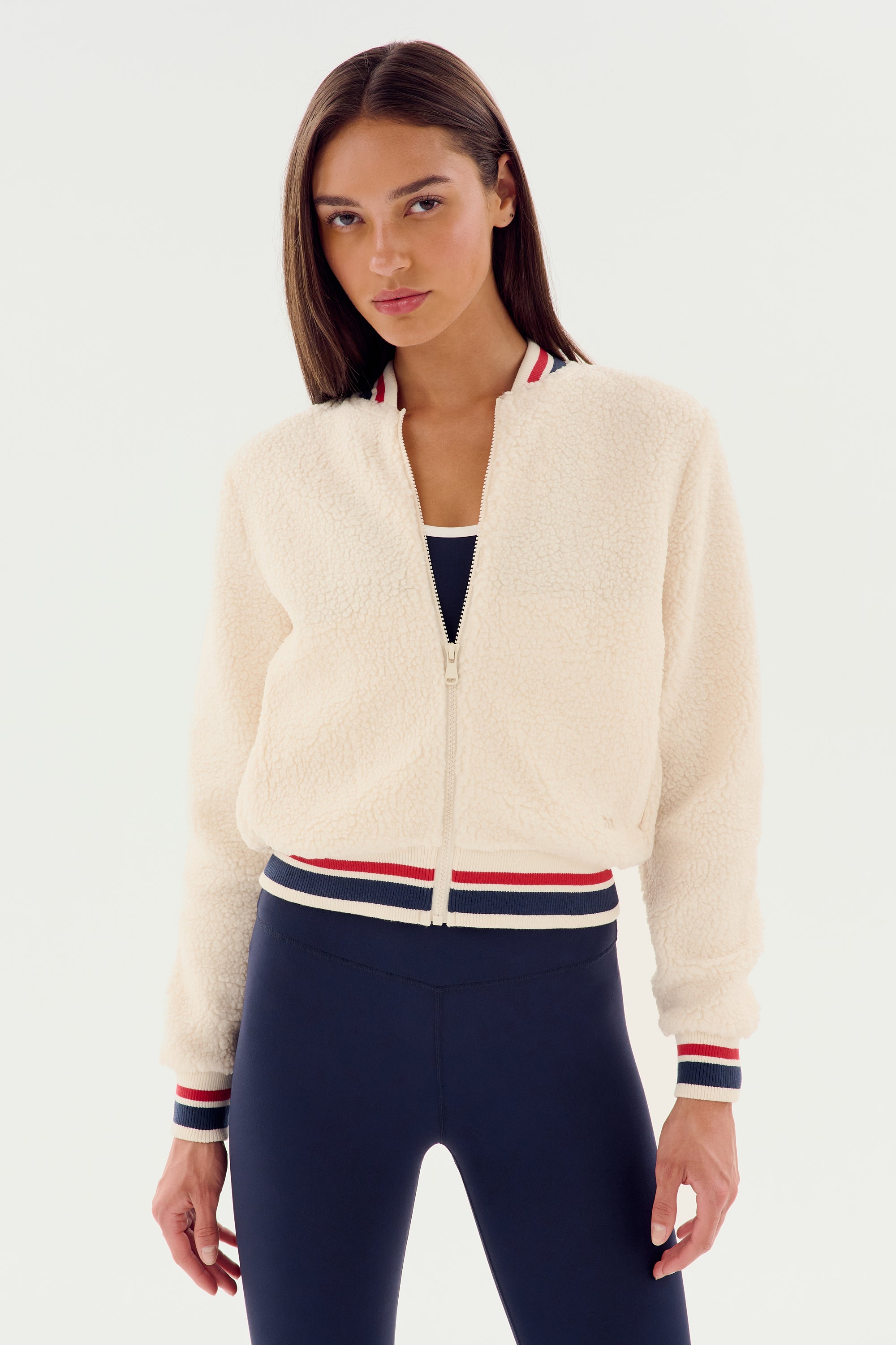 A person is wearing the Willa Sherpa Jacket - Creme by SPLITS59, featuring a zip-up design with red, white, and blue striped collar and cuffs, perfect for winter workouts, paired with black pants.