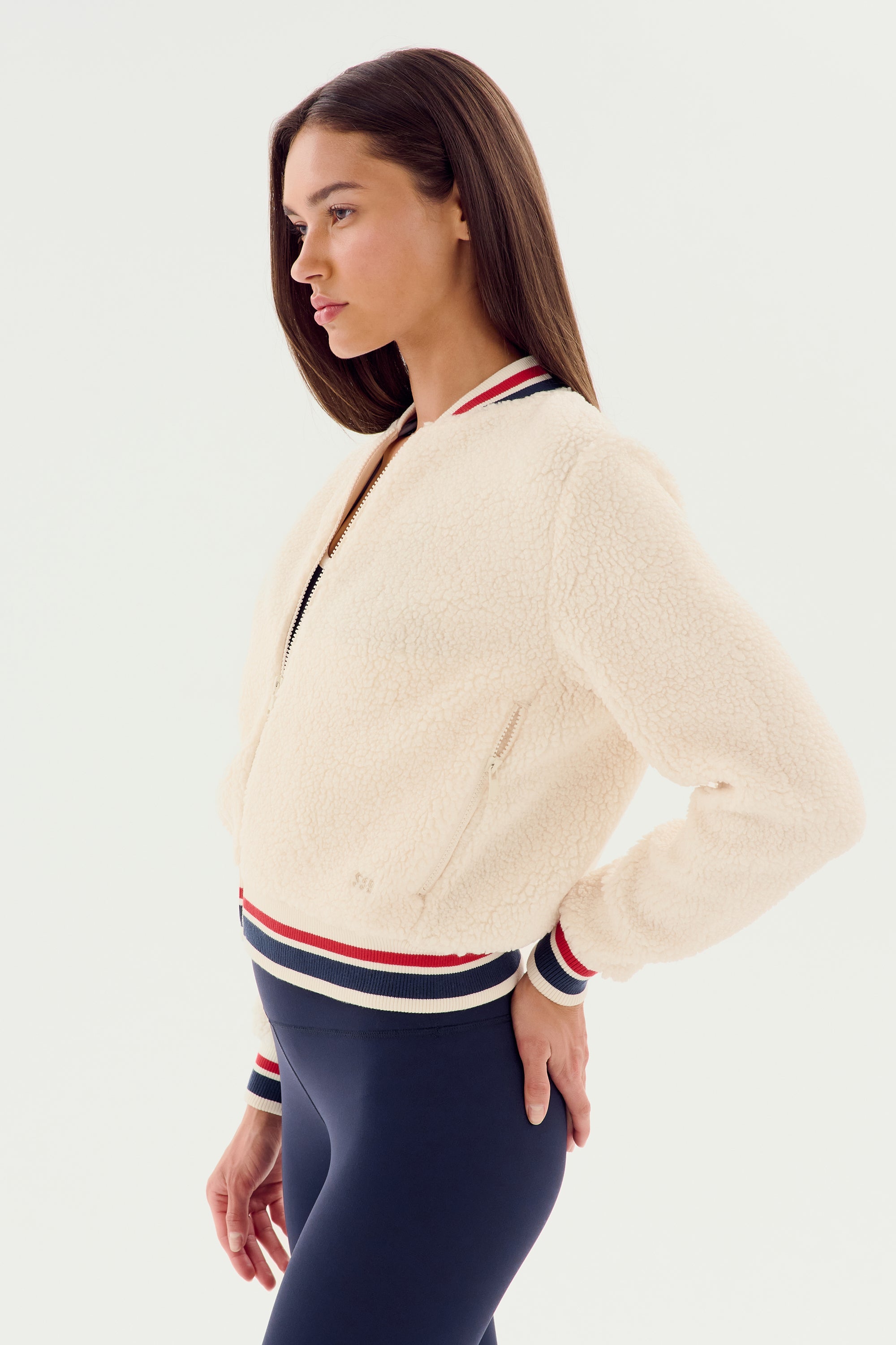 A woman stands in profile against a white background, wearing the SPLITS59 Willa Sherpa Jacket in cream with striped collar and cuffs, paired with navy leggings for a cozy sportswear look.