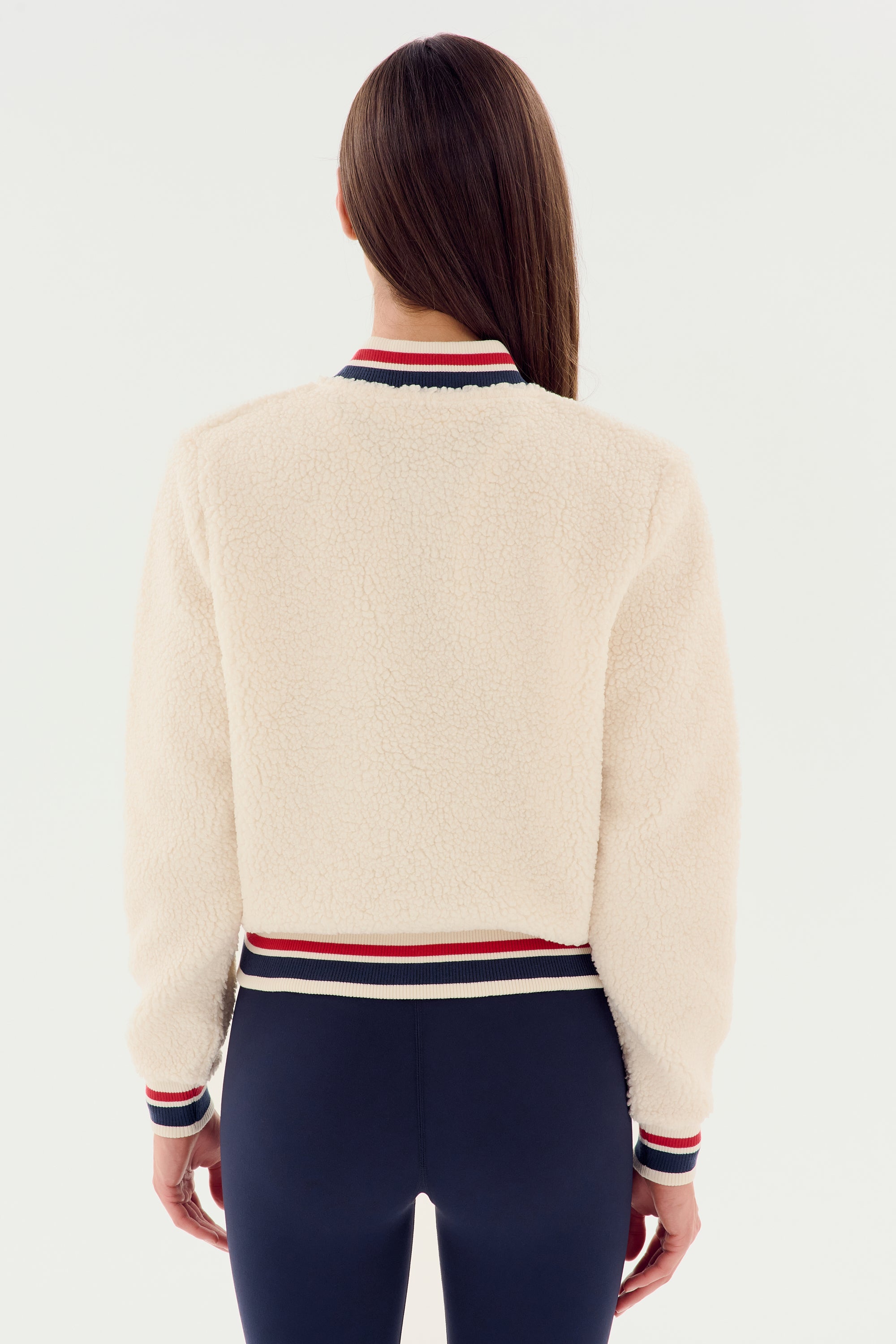 A person with long brown hair wears the SPLITS59 Willa Sherpa Jacket in cream, featuring striped cuffs and collar, paired with black fitted pants.