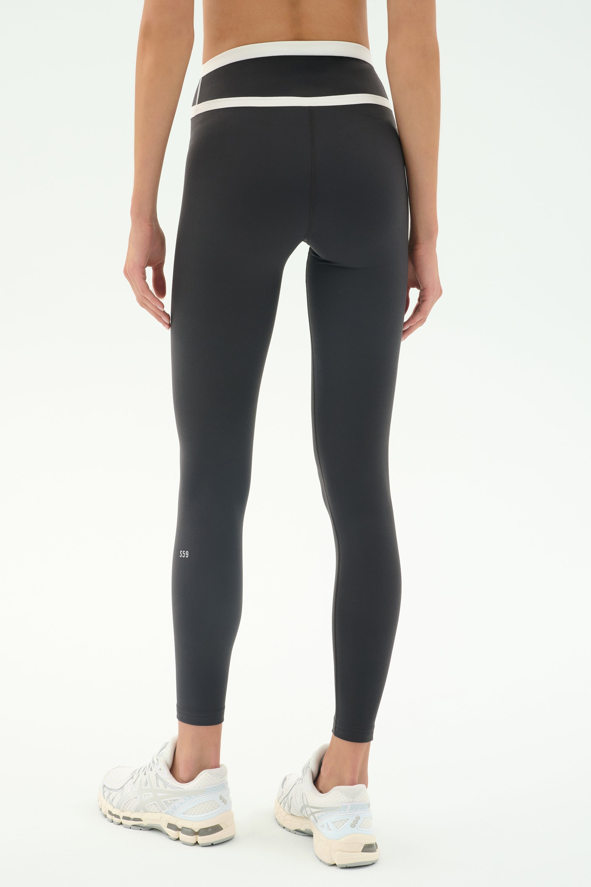 A person wearing SPLITS59 Mason High Waist Rigor Full Length leggings in Graphite/White, paired with white sneakers, stands facing away from the camera against a plain background.