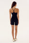 A person with long hair is standing and facing away, wearing a black body-hugging Airweight 6” Short Jumpsuit - Black from SPLITS59 and white sneakers.