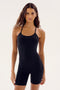 A woman with long hair is wearing a black, body-hugging Airweight 6” Short Jumpsuit - Black from SPLITS59, standing against a plain white background.