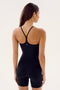 A woman with long hair faces away, wearing a body-hugging black Airweight 6” Short Jumpsuit - Black by SPLITS59, perfect for hot yoga, against a plain background.