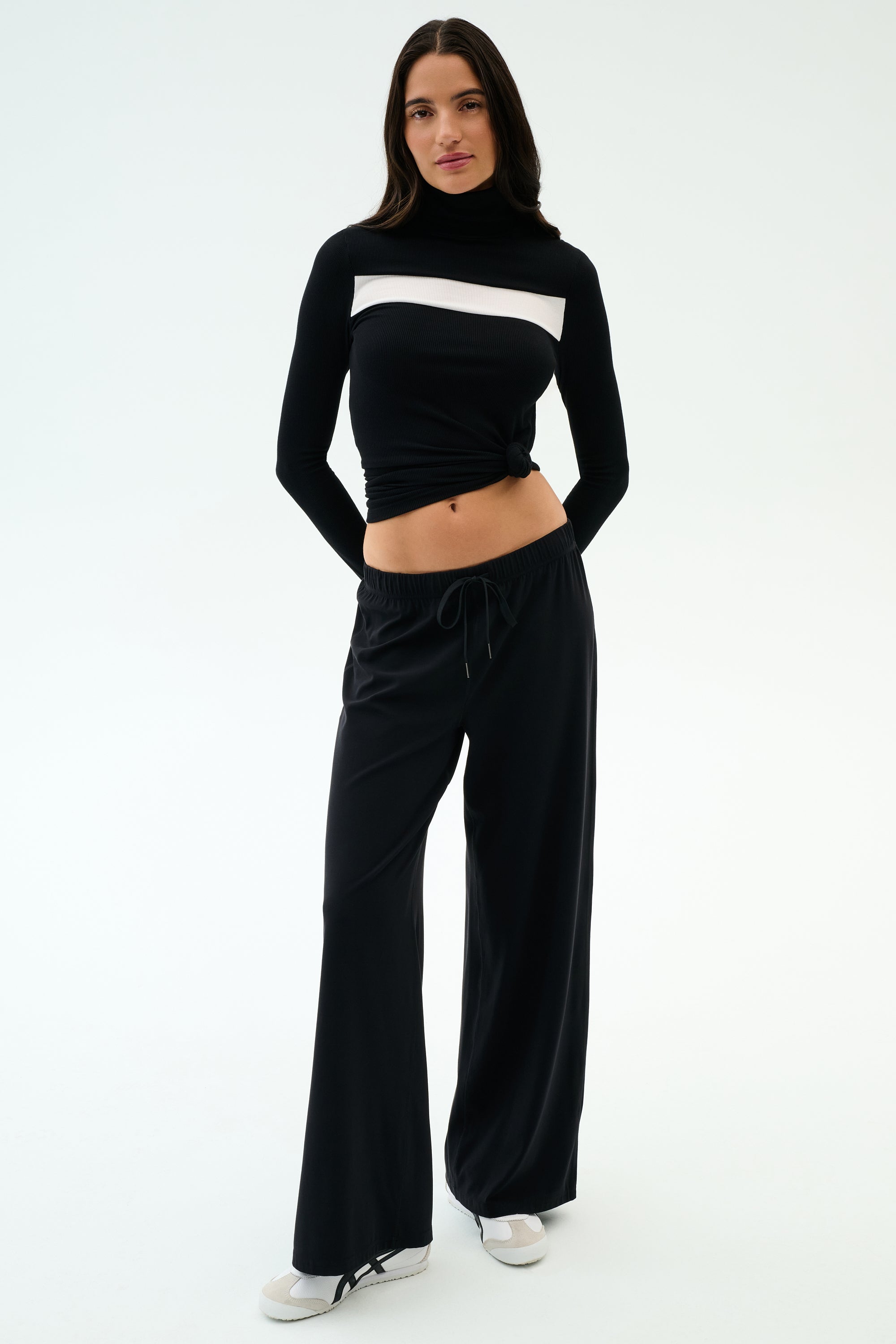 A woman stands facing forward against a plain background wearing SPLITS59's Brynn Airweight Pant in black, paired with a black nylon long-sleeve turtleneck featuring a white horizontal stripe and white shoes.
