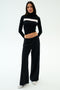 A woman stands facing forward against a plain background wearing SPLITS59's Brynn Airweight Pant in black, paired with a black nylon long-sleeve turtleneck featuring a white horizontal stripe and white shoes.