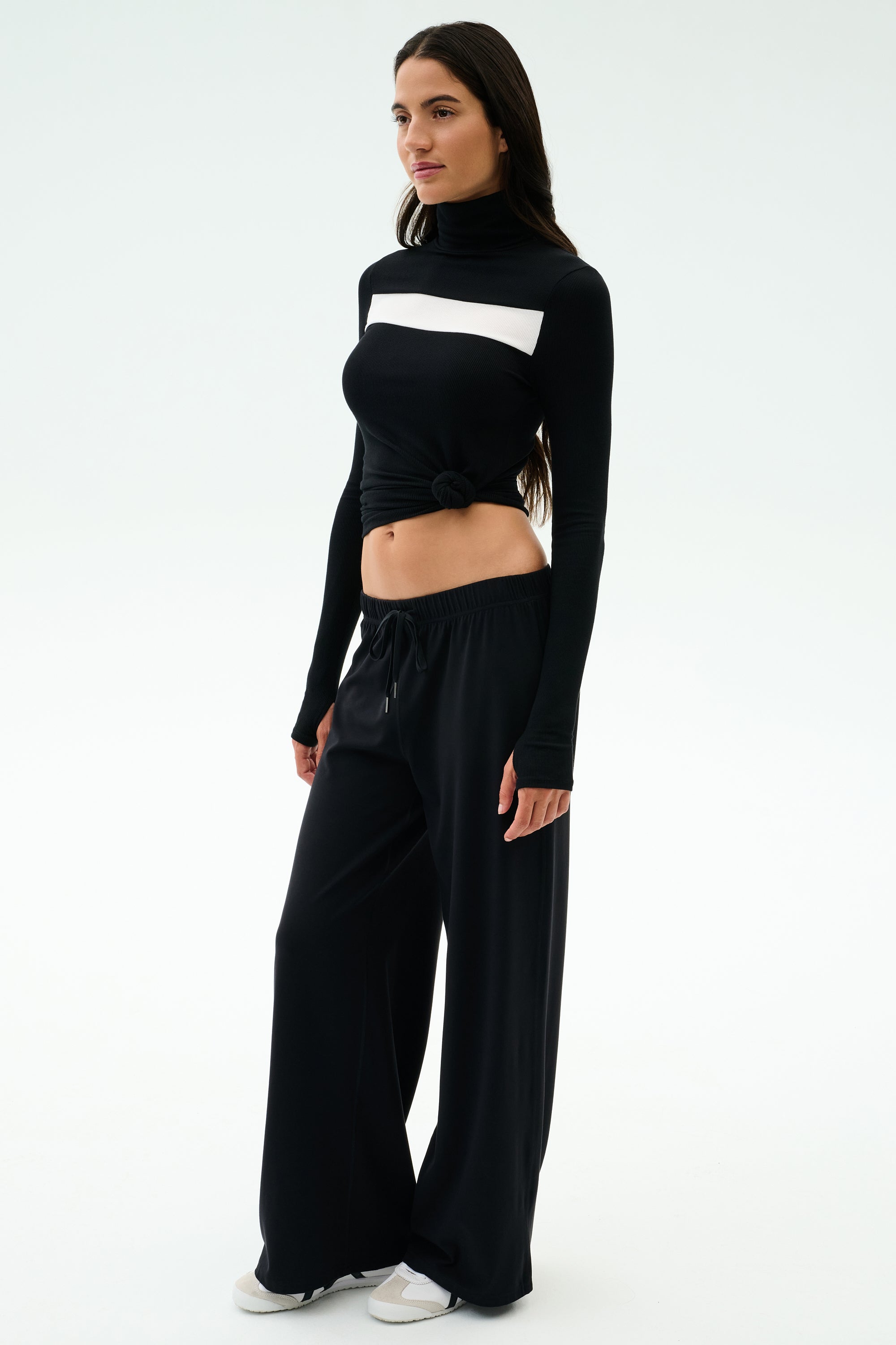 A woman stands against a plain background, wearing the SPLITS59 Brynn Airweight Pant in black, paired with a black and white long-sleeve spandex front-knot top and white sneakers.