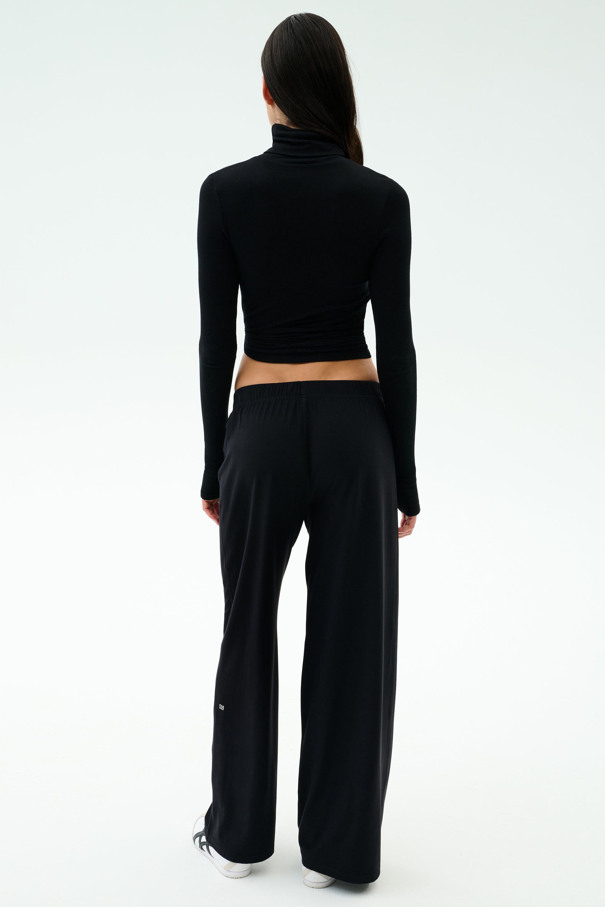 A person faces away, wearing SPLITS59's Brynn Airweight Pant in black, paired with a black spandex long-sleeve turtleneck and white sneakers against a plain light background.