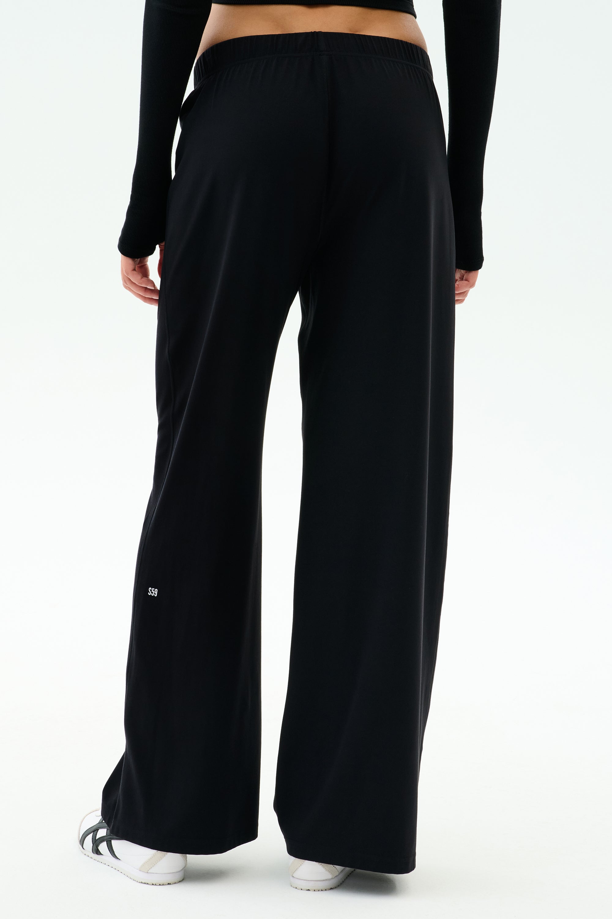 Shown from the back against a plain white background, a person wears SPLITS59 Brynn Airweight Pant in black with a black long-sleeve spandex top and white shoes.