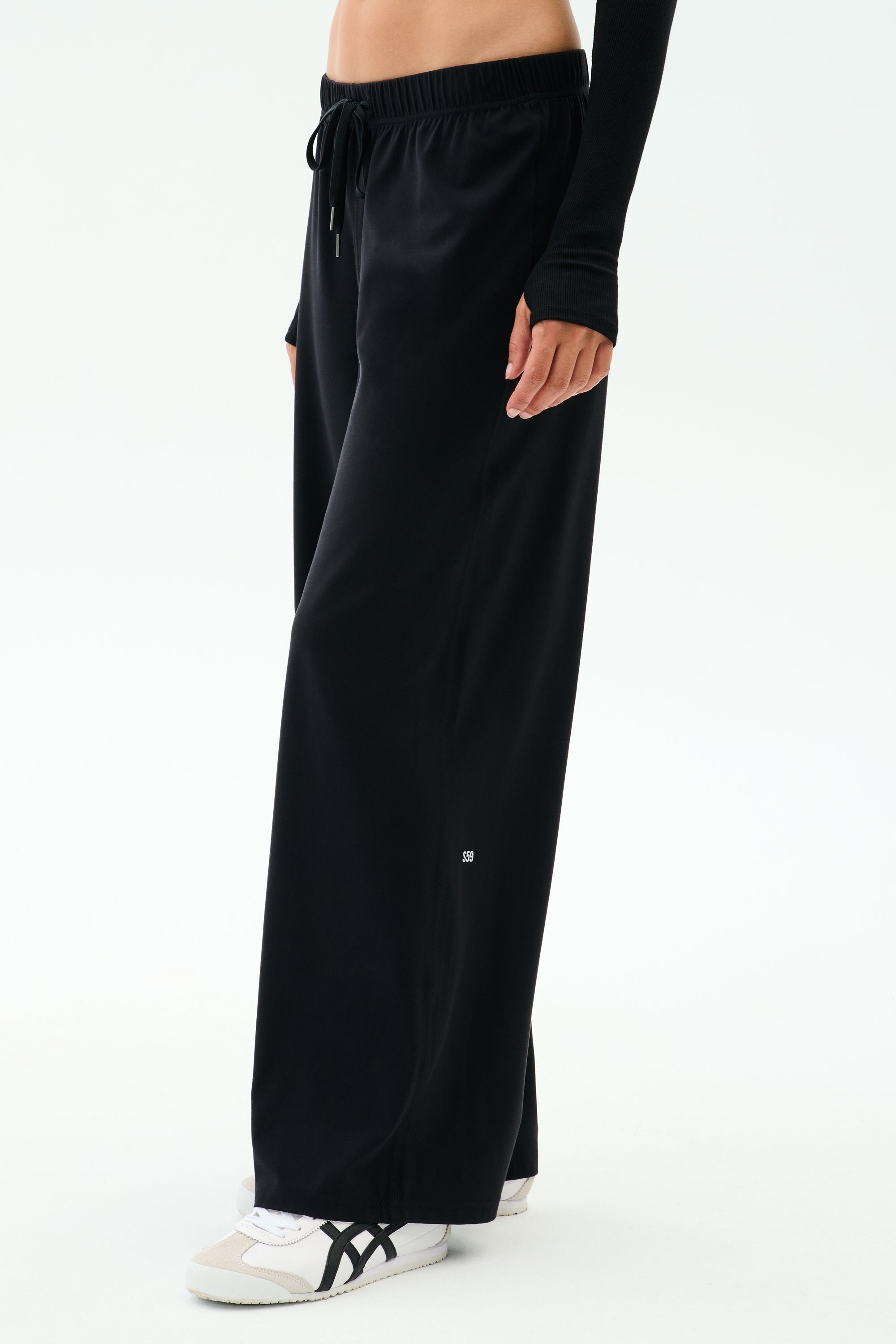 A person wears SPLITS59 Brynn Airweight Pant in black, styled with white sneakers featuring black stripes and a long-sleeve black top, standing against a plain light background.