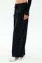 A person wears SPLITS59 Brynn Airweight Pant in black, styled with white sneakers featuring black stripes and a long-sleeve black top, standing against a plain light background.