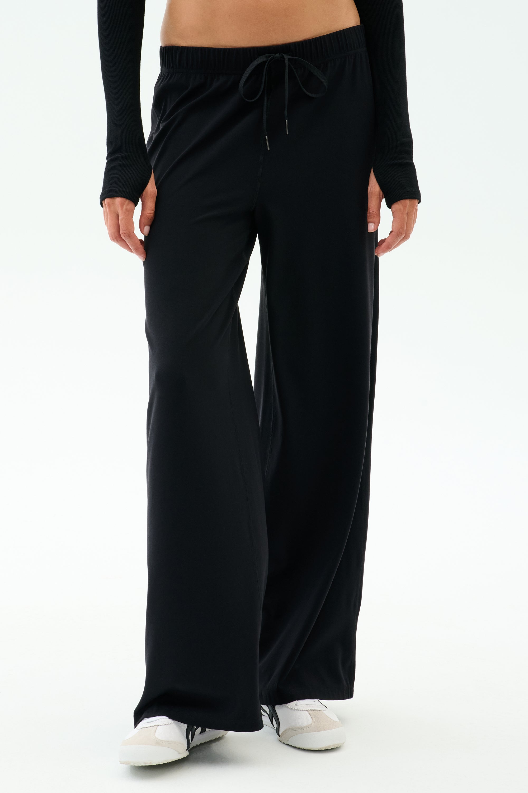 Wearing SPLITS59's Brynn Airweight Pant in Black with a black long-sleeve top and white sneakers, the person stands against a plain white background.