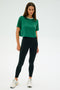 A person wearing the SPLITS59 Djuna Crop Ringer in Arugula/White and black leggings made from an elastane blend poses confidently against a plain backdrop.