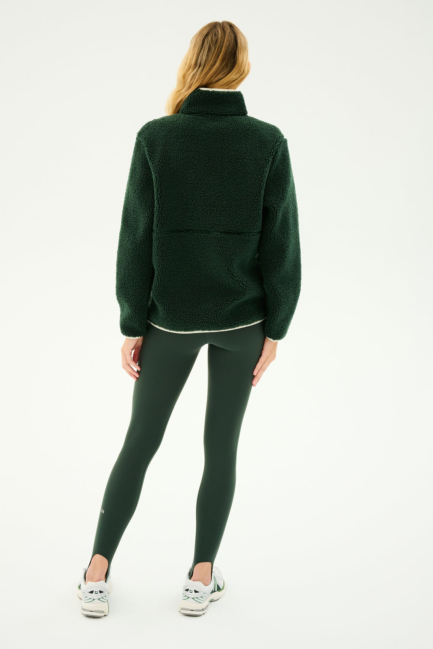 A person with long blonde hair is standing with their back to the camera, sporting the SPLITS59 Libby Sherpa Half Zip in Military/Creme, dark green leggings, and white sneakers, appearing ready for winter workouts.