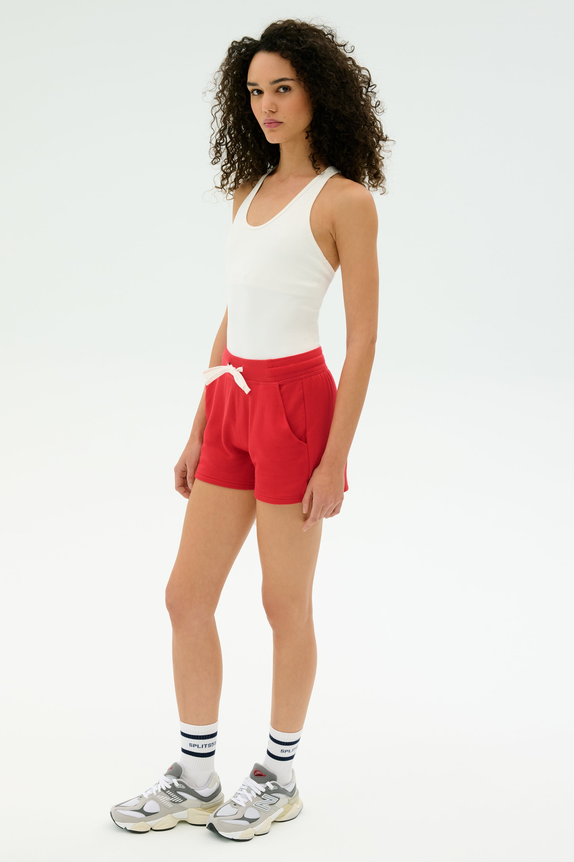 A woman stands on a white background wearing a white tank top, SPLITS59 Reena Sweatshort in Pirate Red, white socks, and gray sneakers.