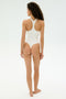 A person with curly hair stands barefoot, facing away, wearing the SPLITS59 Suki Airweight Bodysuit in white against a plain, light background.