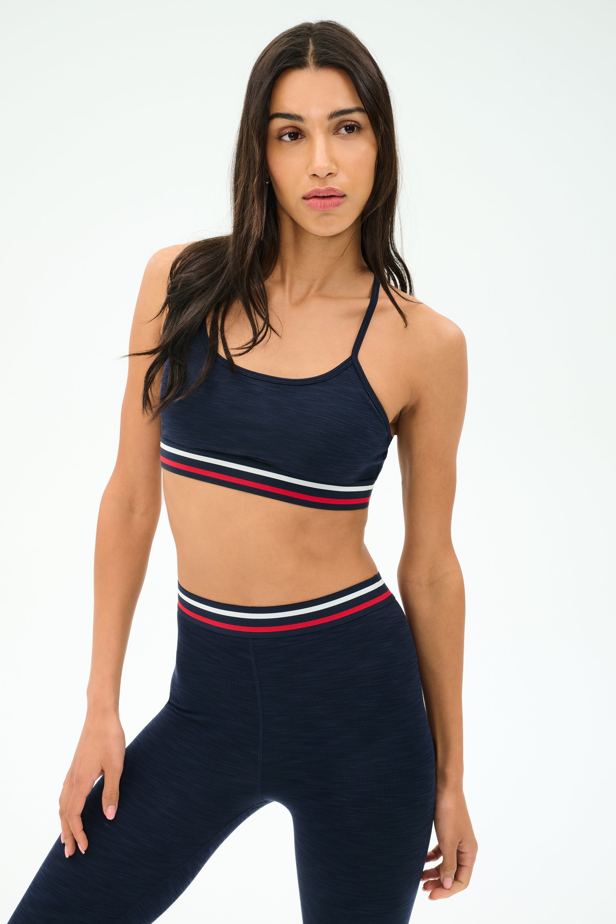 A woman poses confidently in the SPLITS59 Cali Active Rib Bra in Heather Indigo, featuring red, white, and blue striped bands—a supportive choice for your next workout.