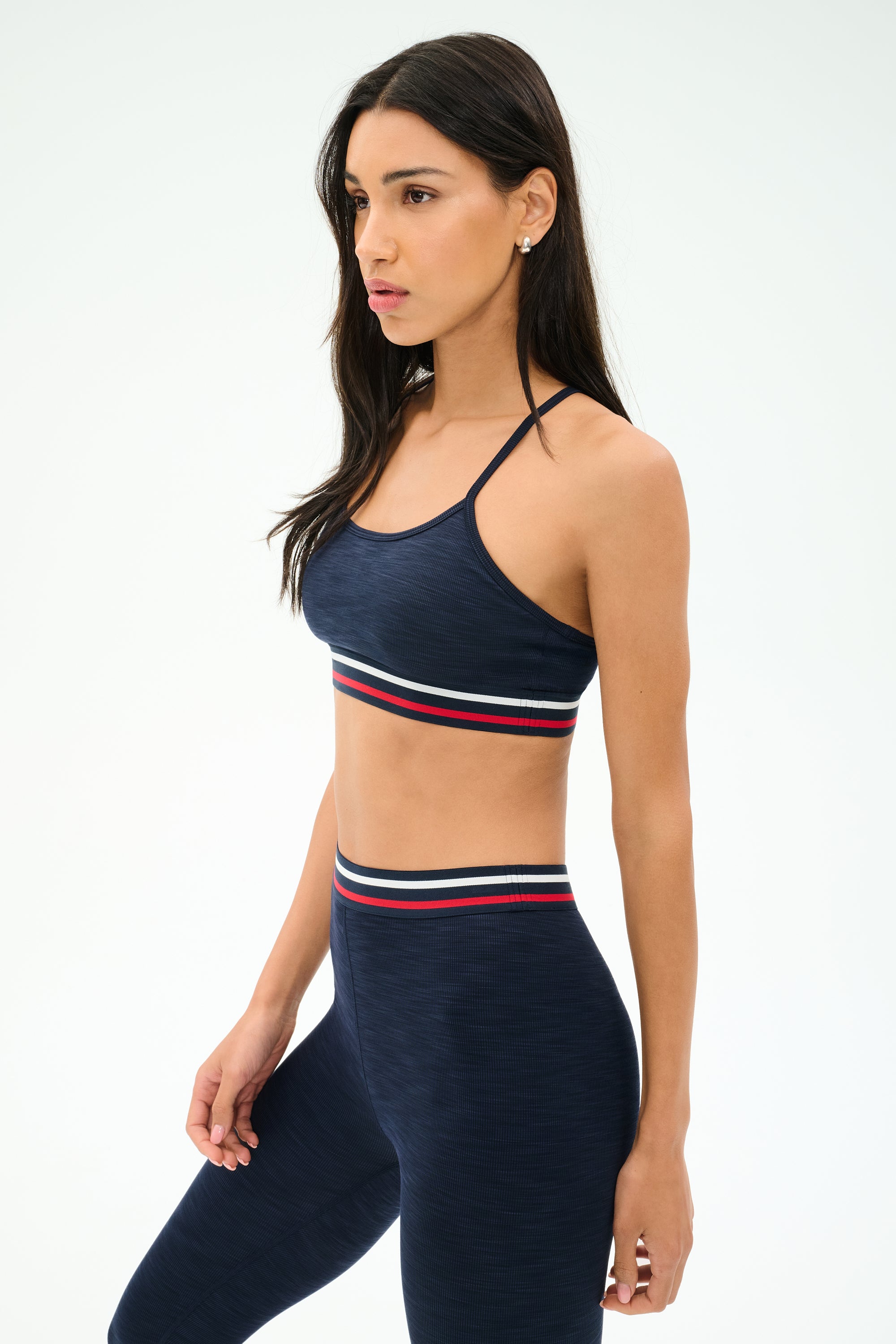 A woman wears the SPLITS59 Cali Active Rib Bra in Heather Indigo with matching leggings featuring red, white, and green striped bands, standing against a plain white background.